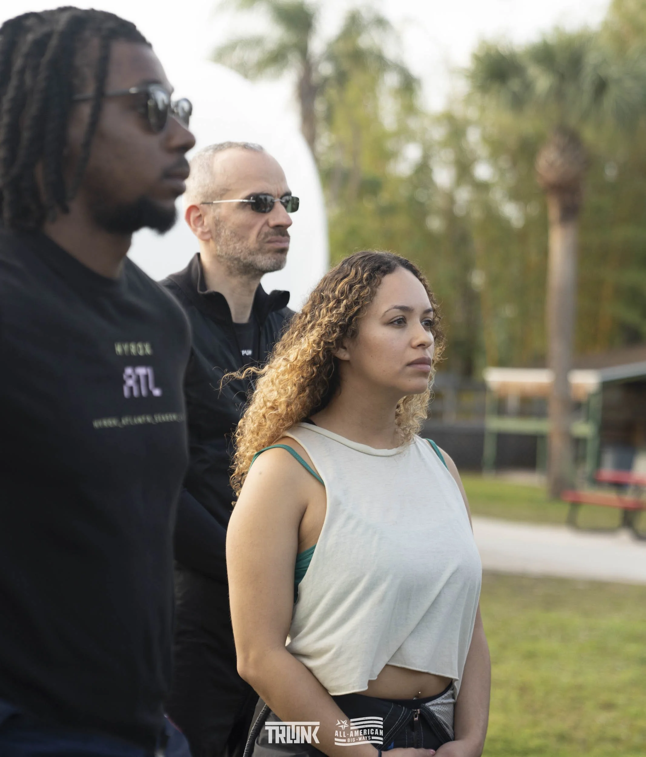 Three people standing outdoors, some trees and a park bench in the background, all wearing serious expressions and sunglasses. The person in the foreground is a woman with curly blonde hair, wearing a beige sleeveless top.