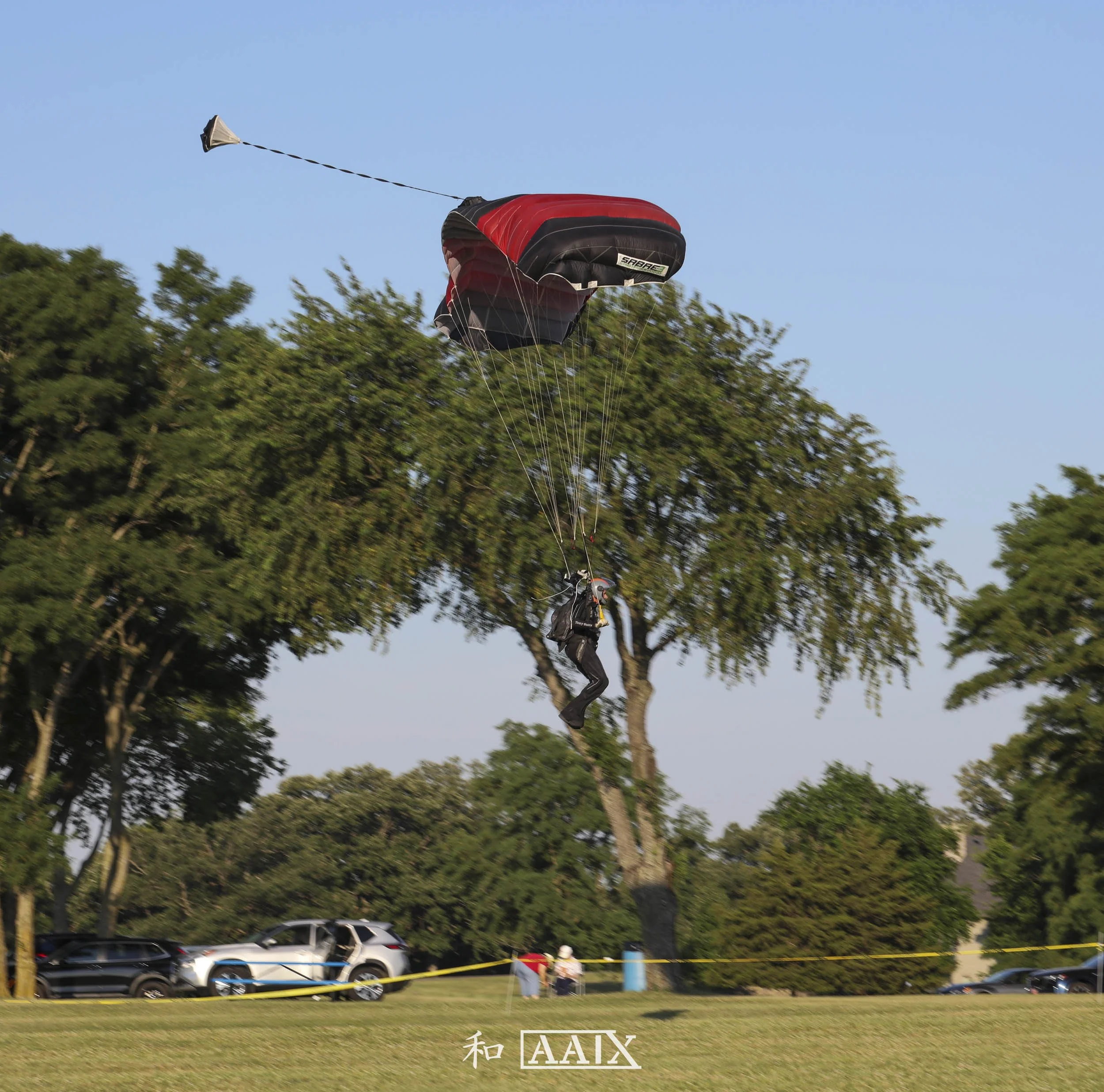 A person in a harness and helmet mid-air during a paragliding jump, descending over a park with trees and parked cars in the background on a clear day.