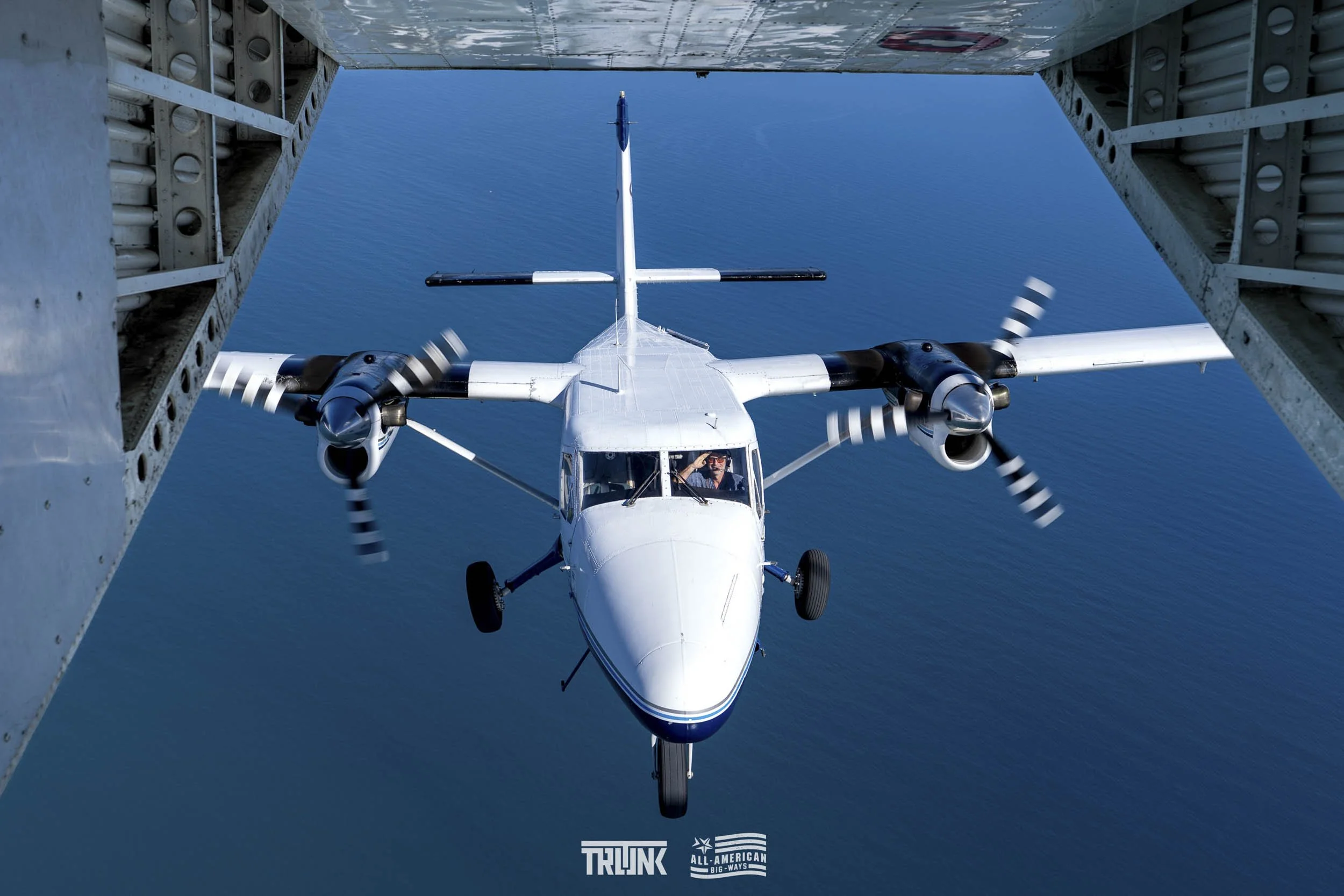 An aircraft flying in formation near an aircraft carrier above the ocean, with wing and fuselage visible in the foreground.