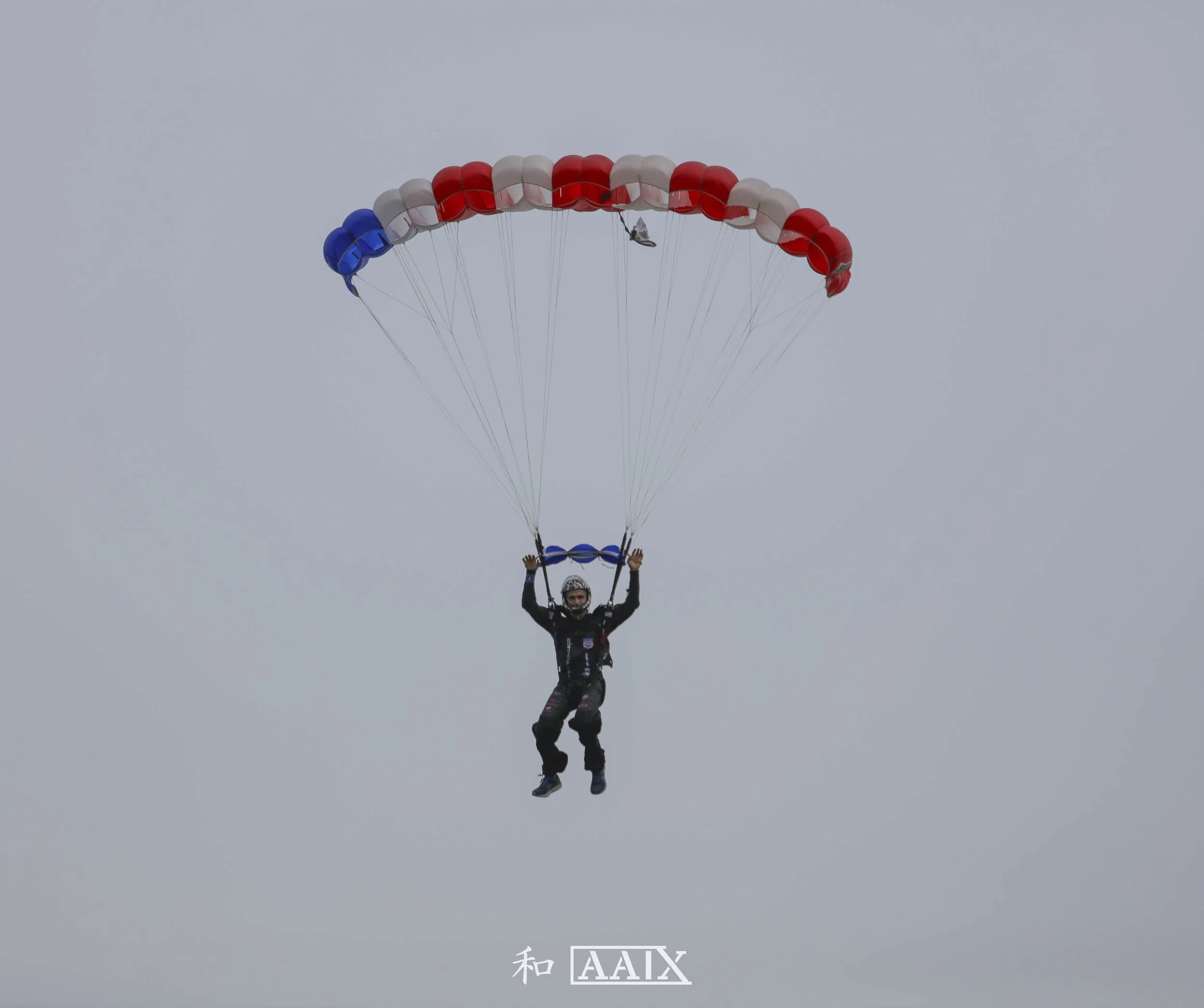 Person parachuting with a canopy displaying the colors of the French flag: blue, white, and red, against a gray sky.
