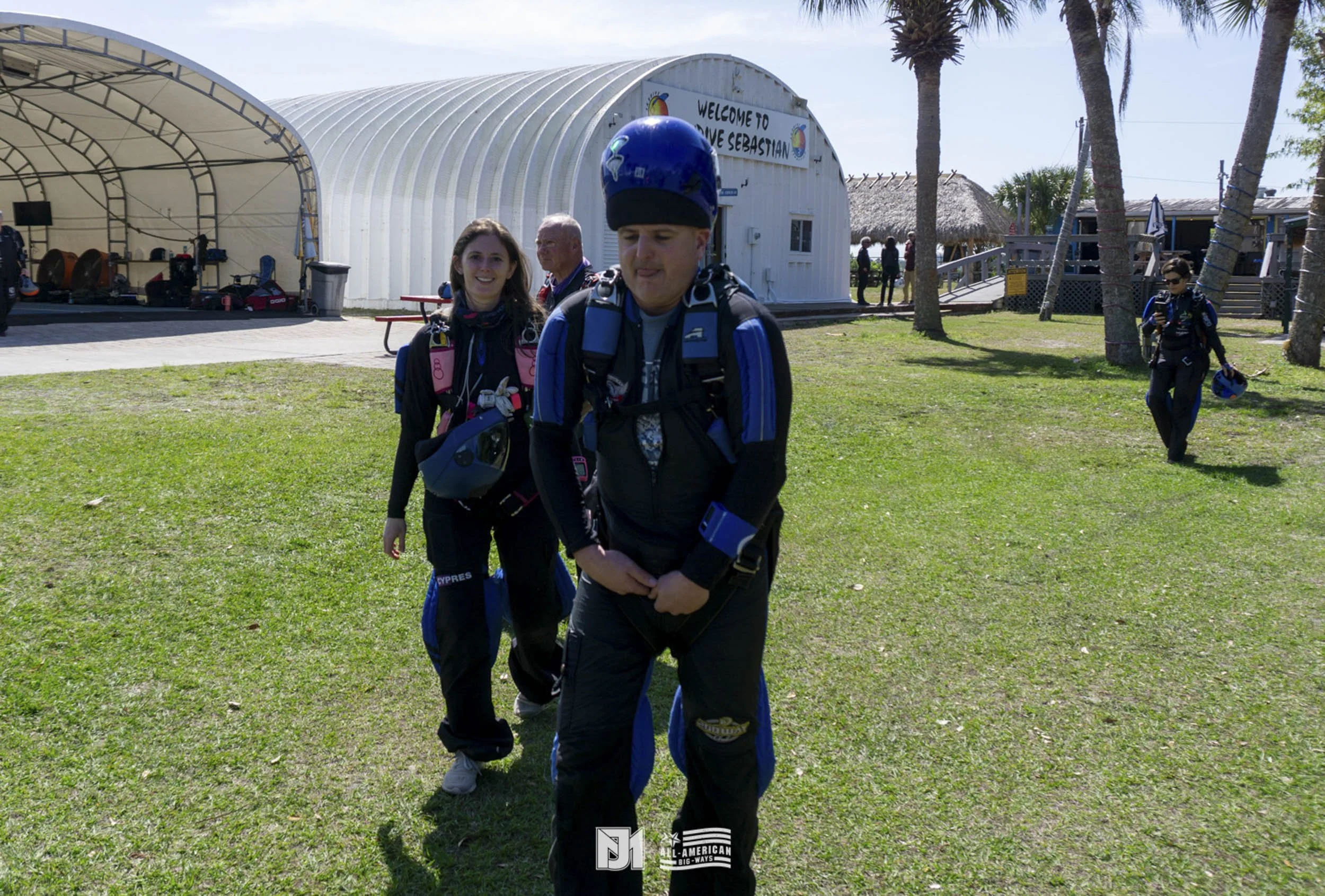 Skydivers walking on grass near a white hangar with a sign that reads 'Welcome to Love Sebastian,' palm trees in the background.