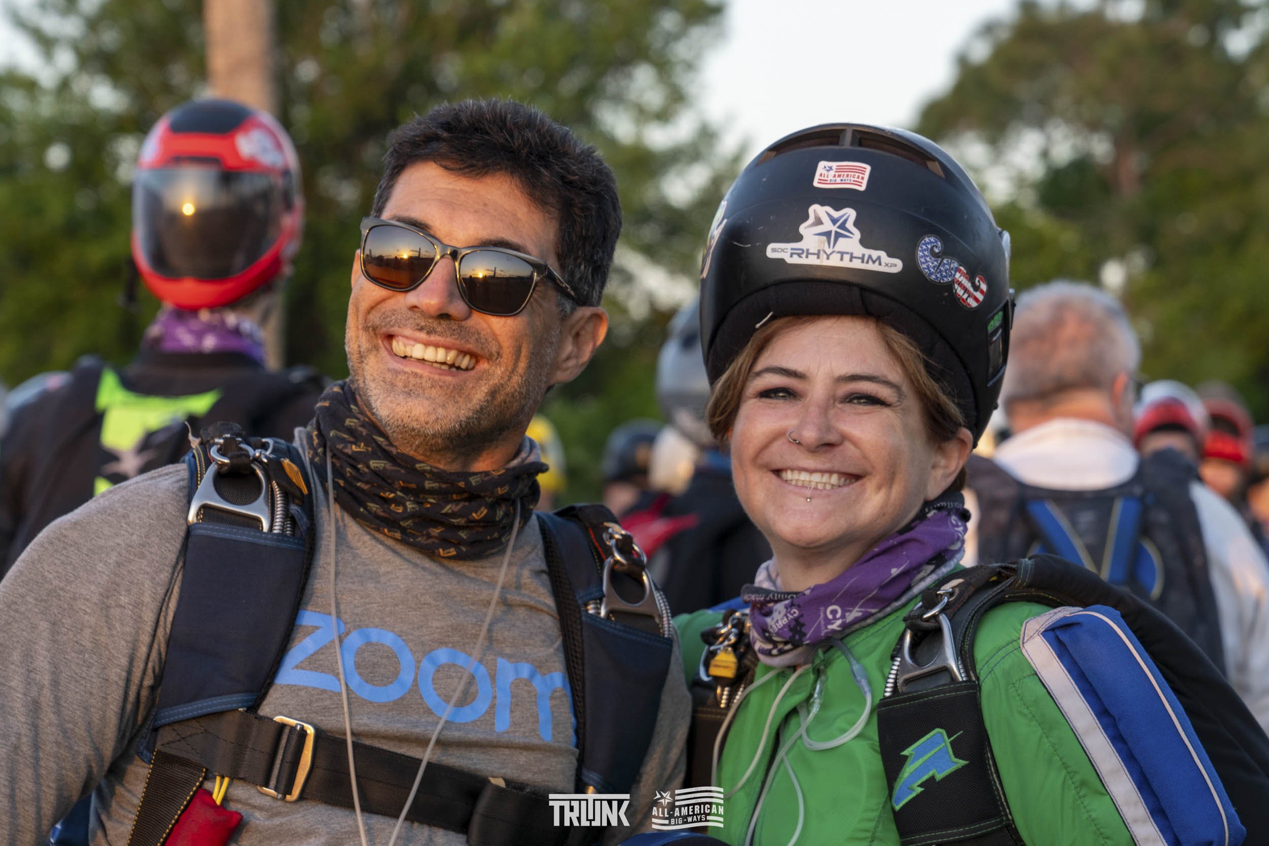 Two smiling people with skydiving gear, one man wearing sunglasses and a gray shirt, and one woman wearing a helmet and green jacket, standing outdoors with a group of skydivers in the background.