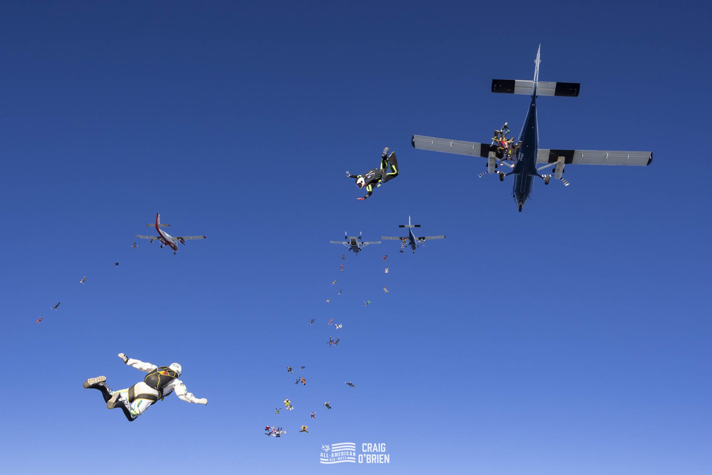 Skydivers and aircrafts mid-air in a formation jump against a clear blue sky, with parachutes deployed.
