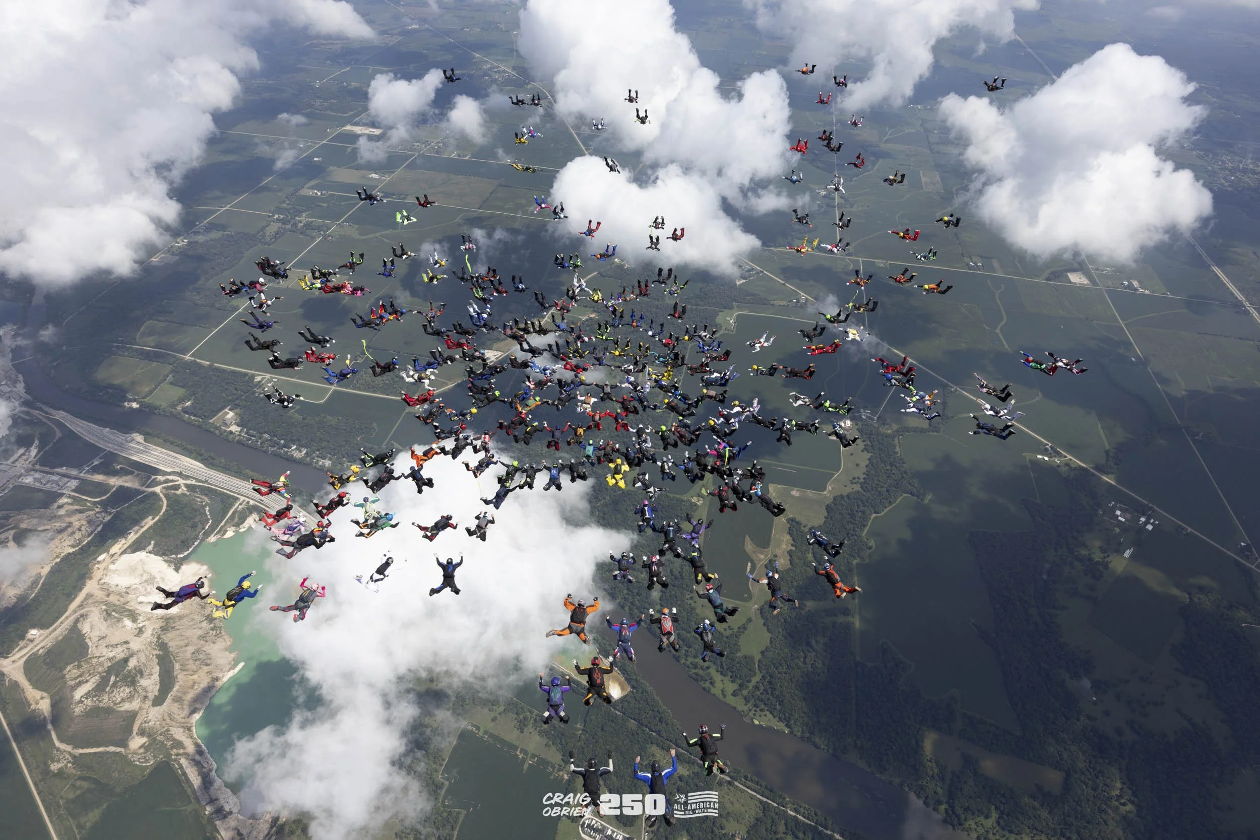 Aerial view of a skydiving formation with numerous skydivers in colorful suits in free fall over a landscape with rivers, farmland, and clouds.