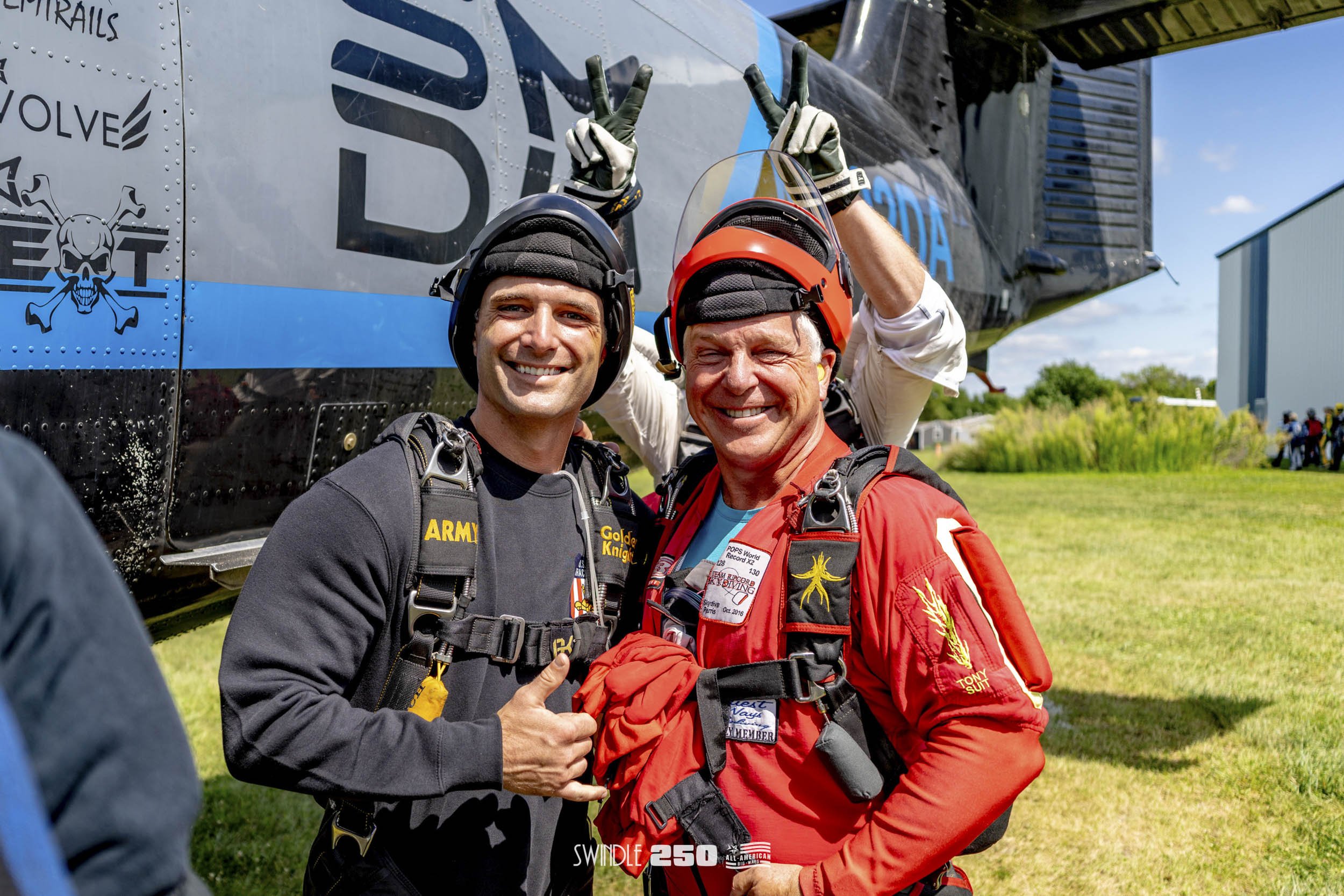 Two skydivers in gear smiling and posing together outdoors near a helicopter. One skydiver is wearing a black jumpsuit with army patches, giving a thumbs-up, and the other is in a red jumpsuit with a helmet, smiling broadly. A third skydiver is parti