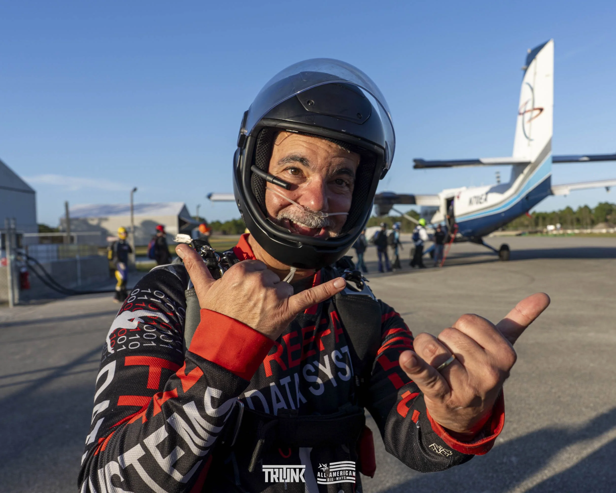 A man in a helmet and racing suit making a shaka gesture in front of an airplane at an airport.