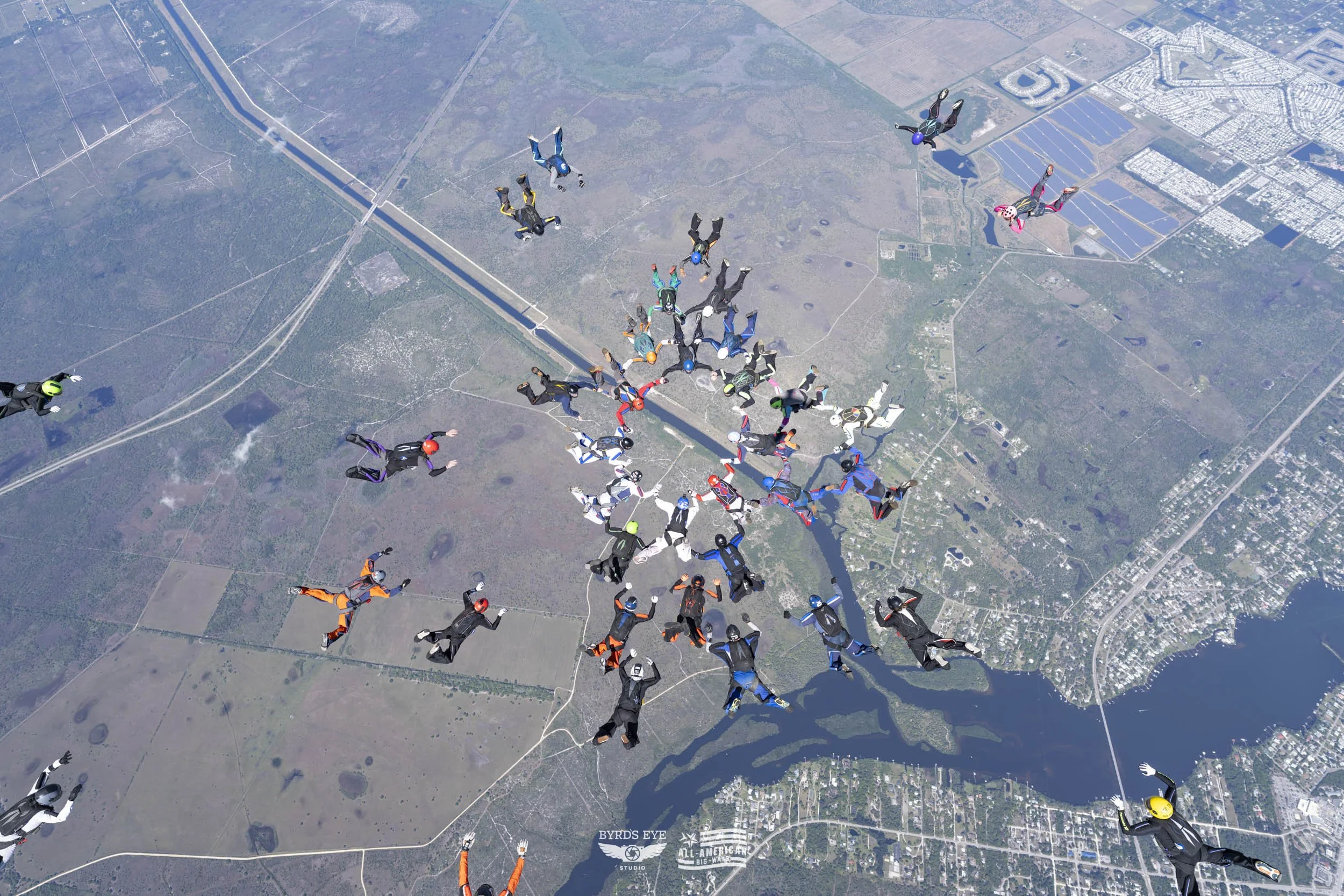 Group of skydivers in free fall over a landscape with lakes, forests, and roads.