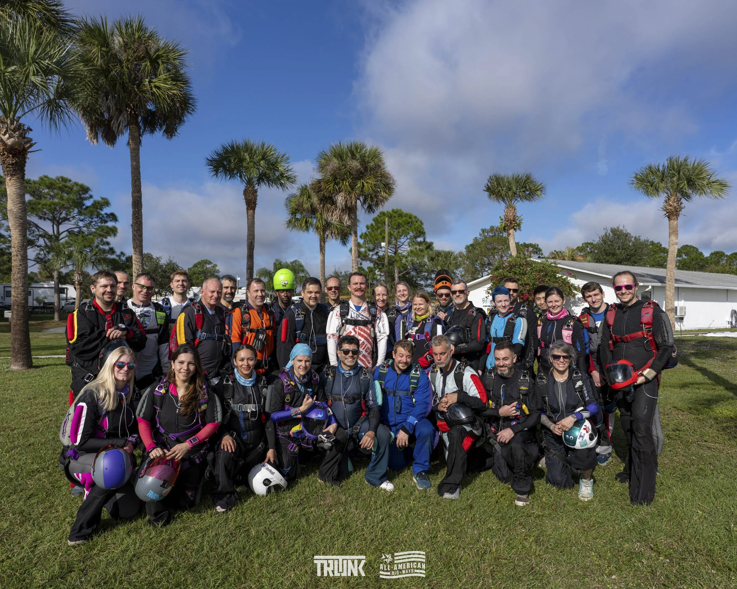 Group of diverse people in outdoor gear, standing and kneeling on grass with palm trees and a building in the background, smiling at the camera