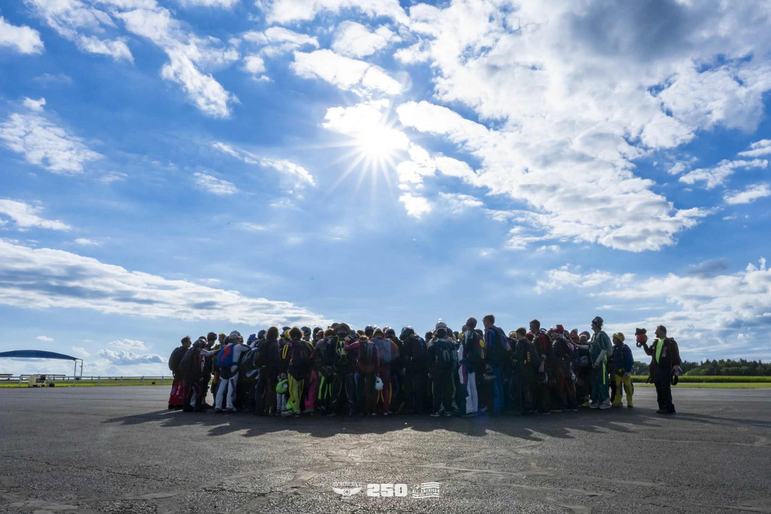 Group of skydivers gathered on tarmac under cloudy sky with the sun shining.