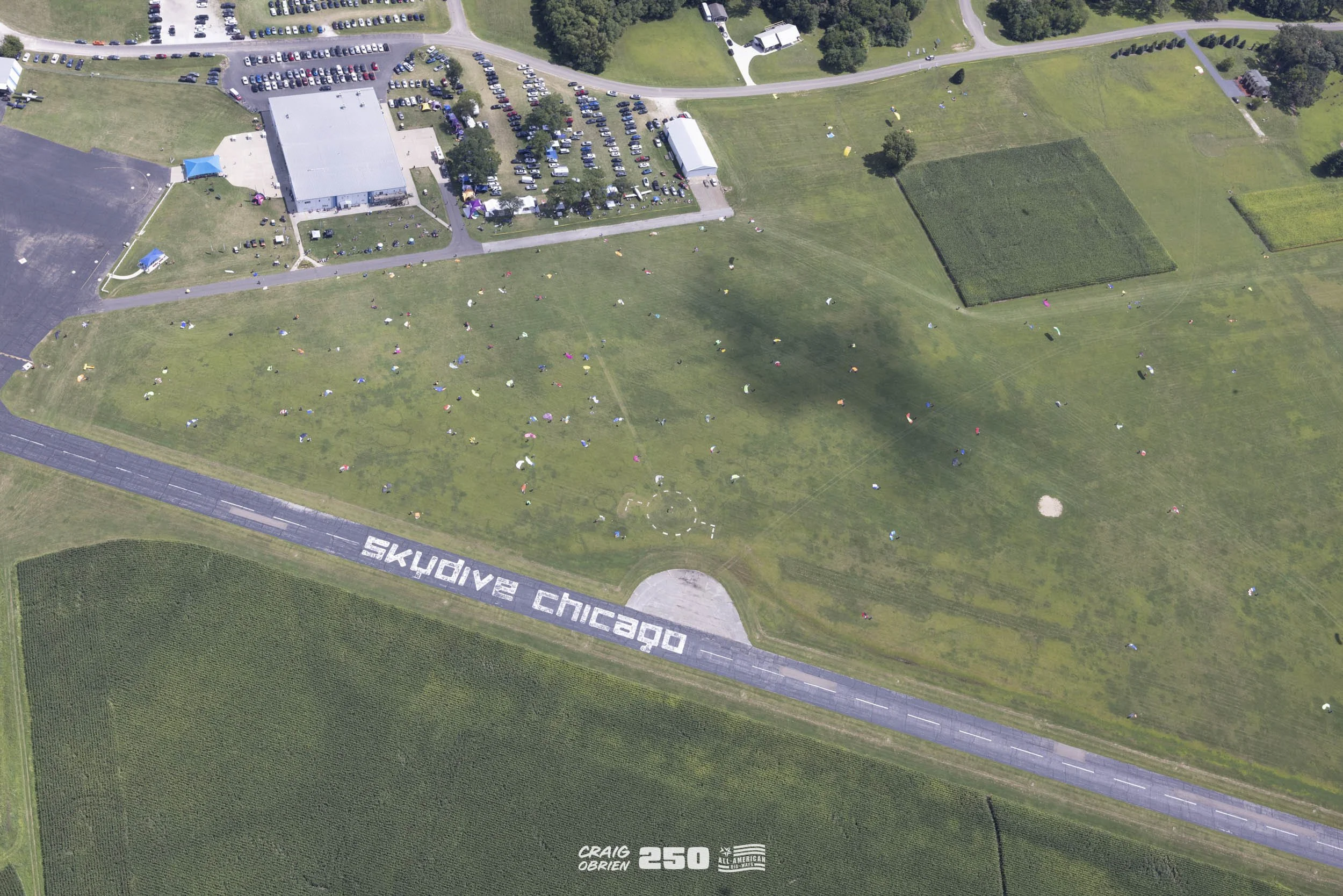 An aerial view of a large green park with many people flying kites. A road with the words 'SKYLOVE CHICAGO' painted on it runs through the scene. There are buildings and parking lots at the top of the image, and fields of crops at the bottom.