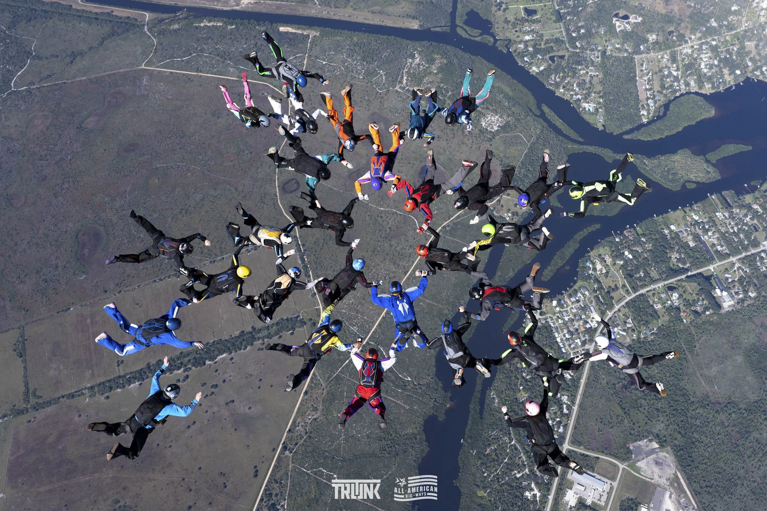 Group of skydivers holding hands in formation during freefall over a landscape with lakes, forests, and towns.