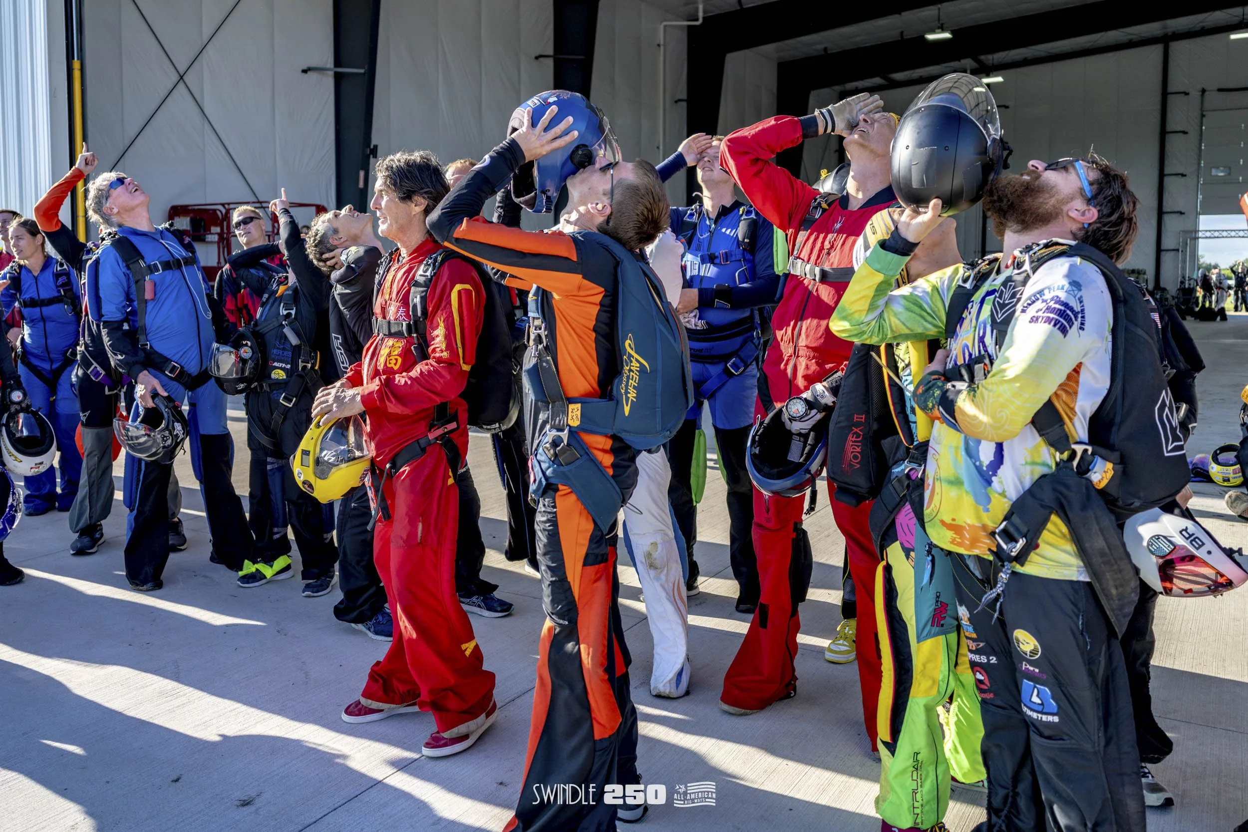 A group of skydivers in jumpsuits and helmets standing together inside a hangar, preparing for a jump.
