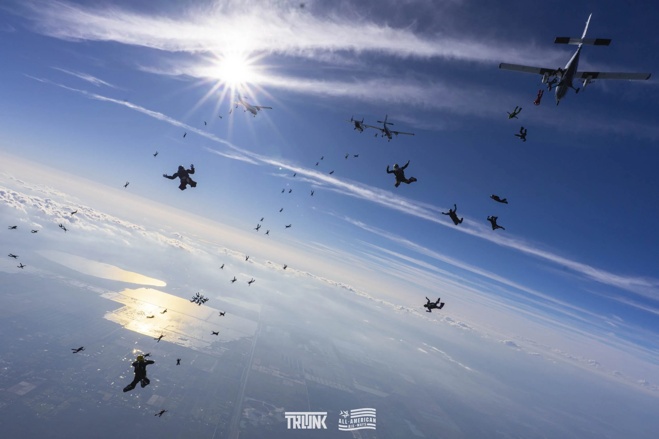 Skydivers jumping from aircraft against a blue sky with some clouds and the sun, with a landscape visible below.