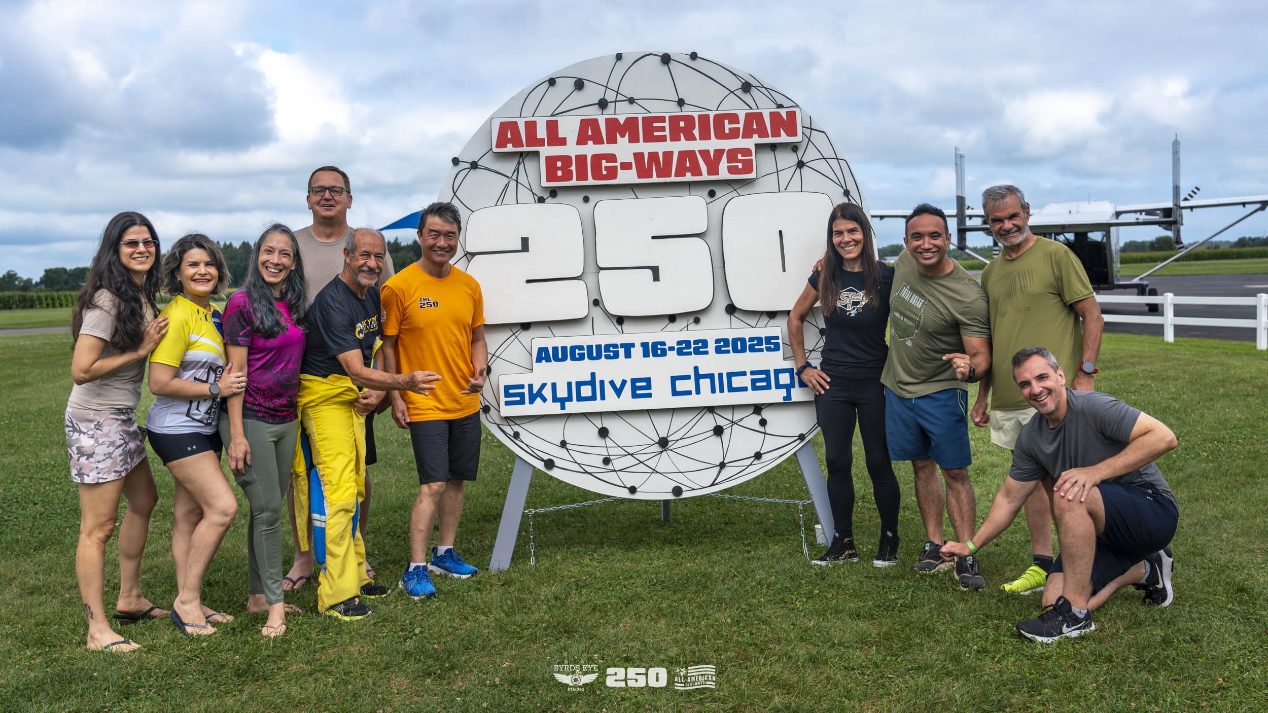 A group of 10 people standing outdoors on a grassy field next to a large sign for skydiving, with an airplane in the background. The sign reads "All American Big-Ways 250, August 16-22, 2025, skydive Chicago." It features a large number 250 and a glo