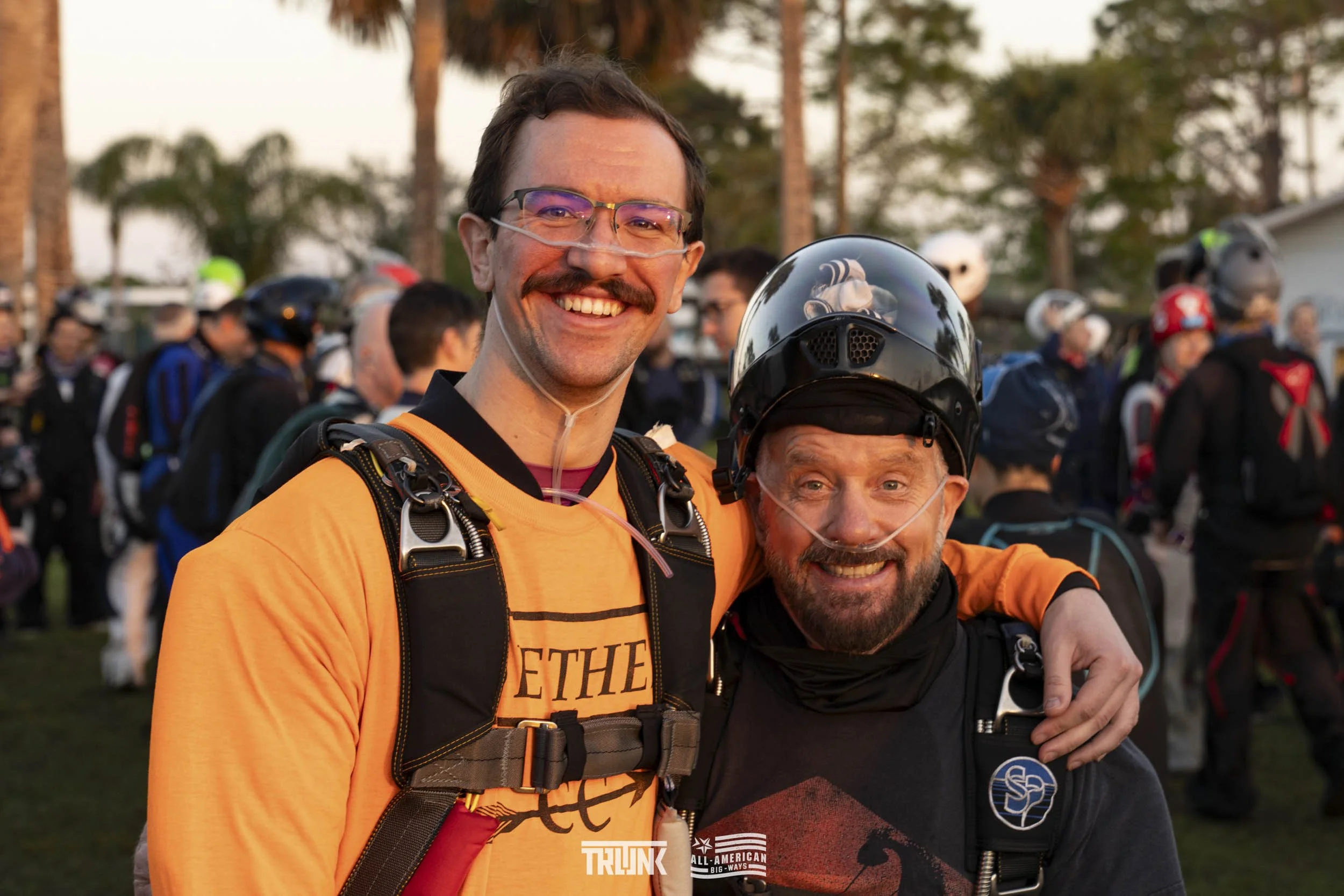Two smiling men at a skydiving event, one wearing an orange shirt and the other with a helmet, surrounded by a group of skydivers in a field with trees in the background.