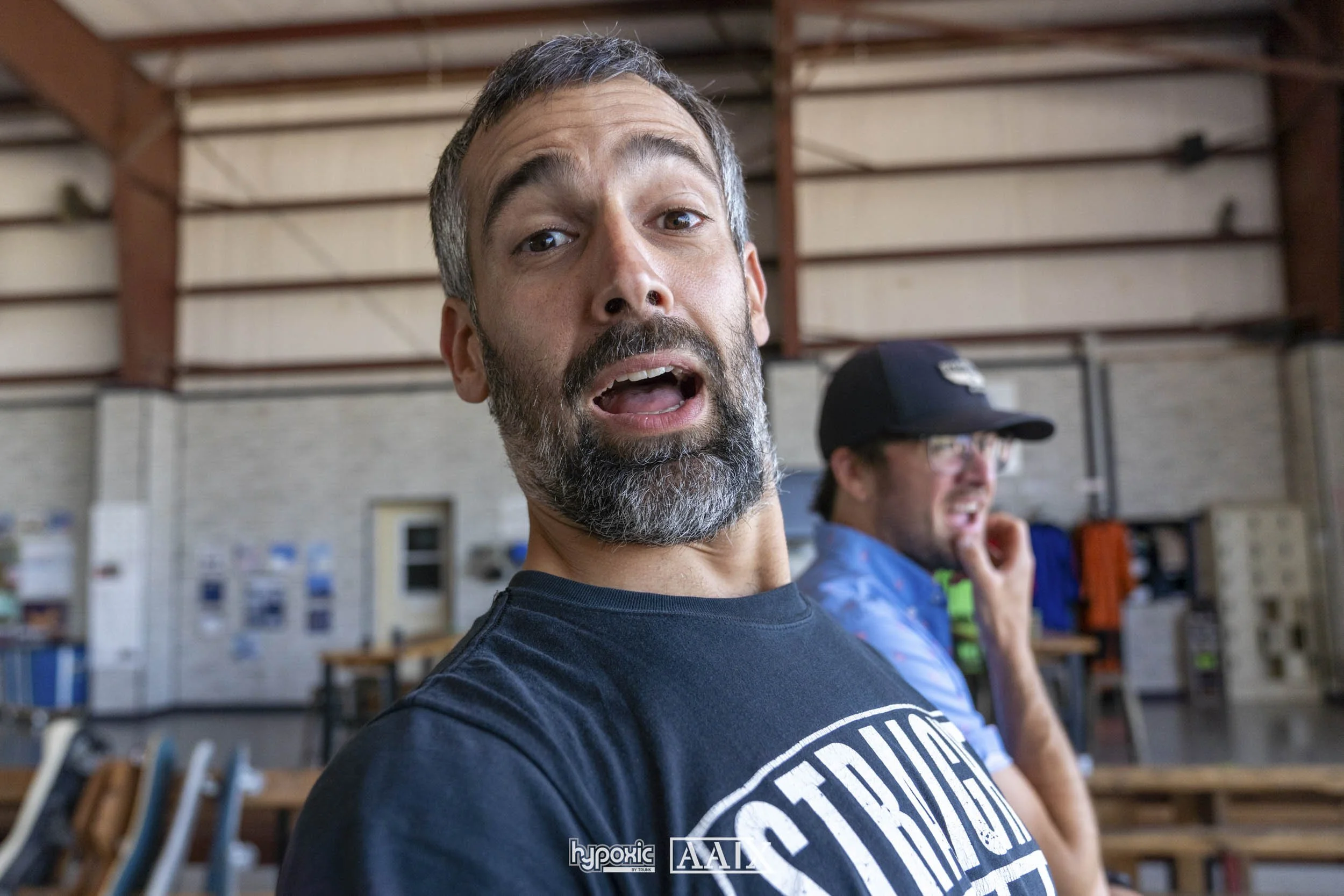A man with gray hair and beard making a surprised or expressive face, wearing a black T-shirt, with another man in a cap and glasses in the background. They are inside a warehouse or workshop.