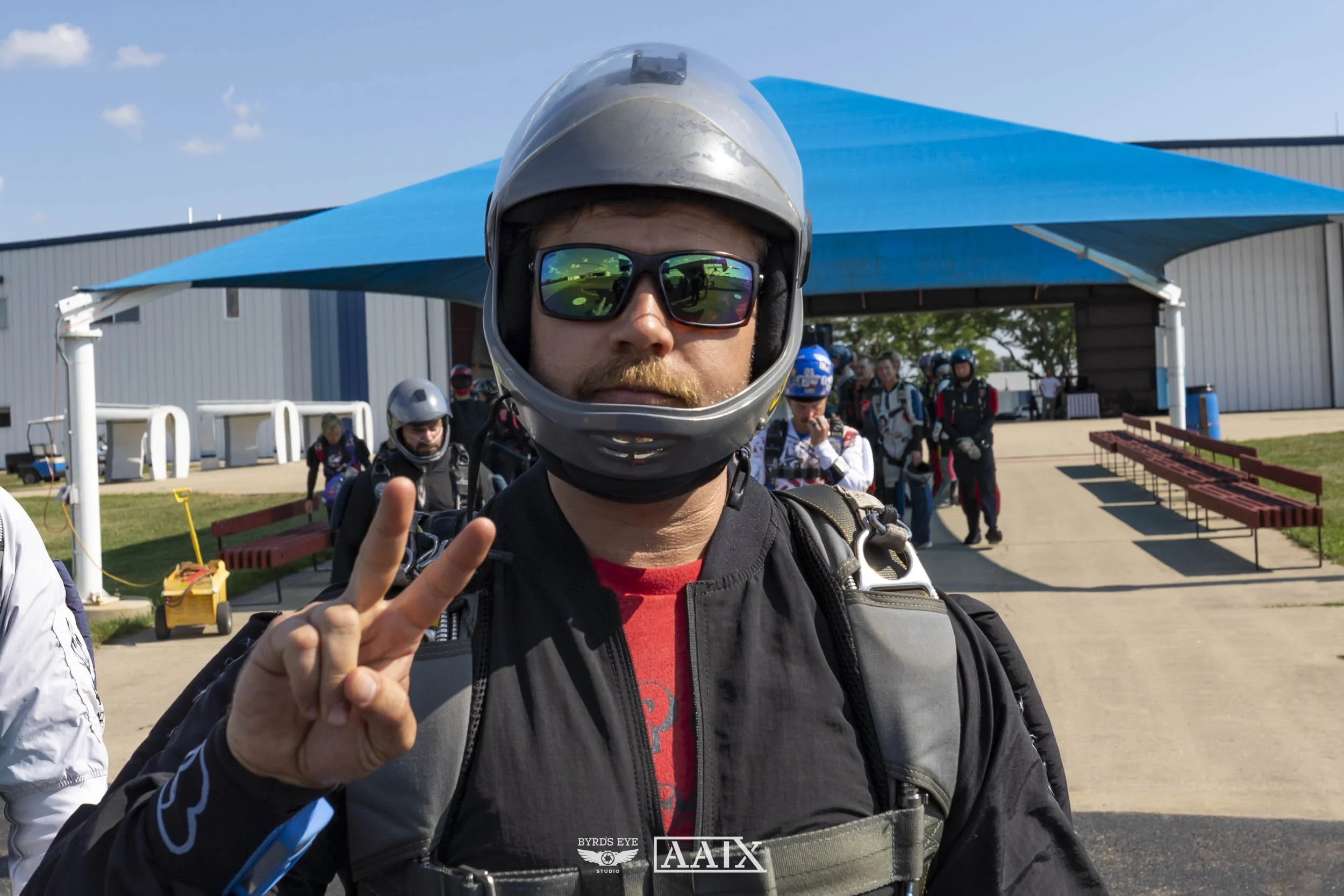 A man in a motorcycle helmet and sunglasses making a peace sign at the camera, with a group of skydivers and a hangar in the background.