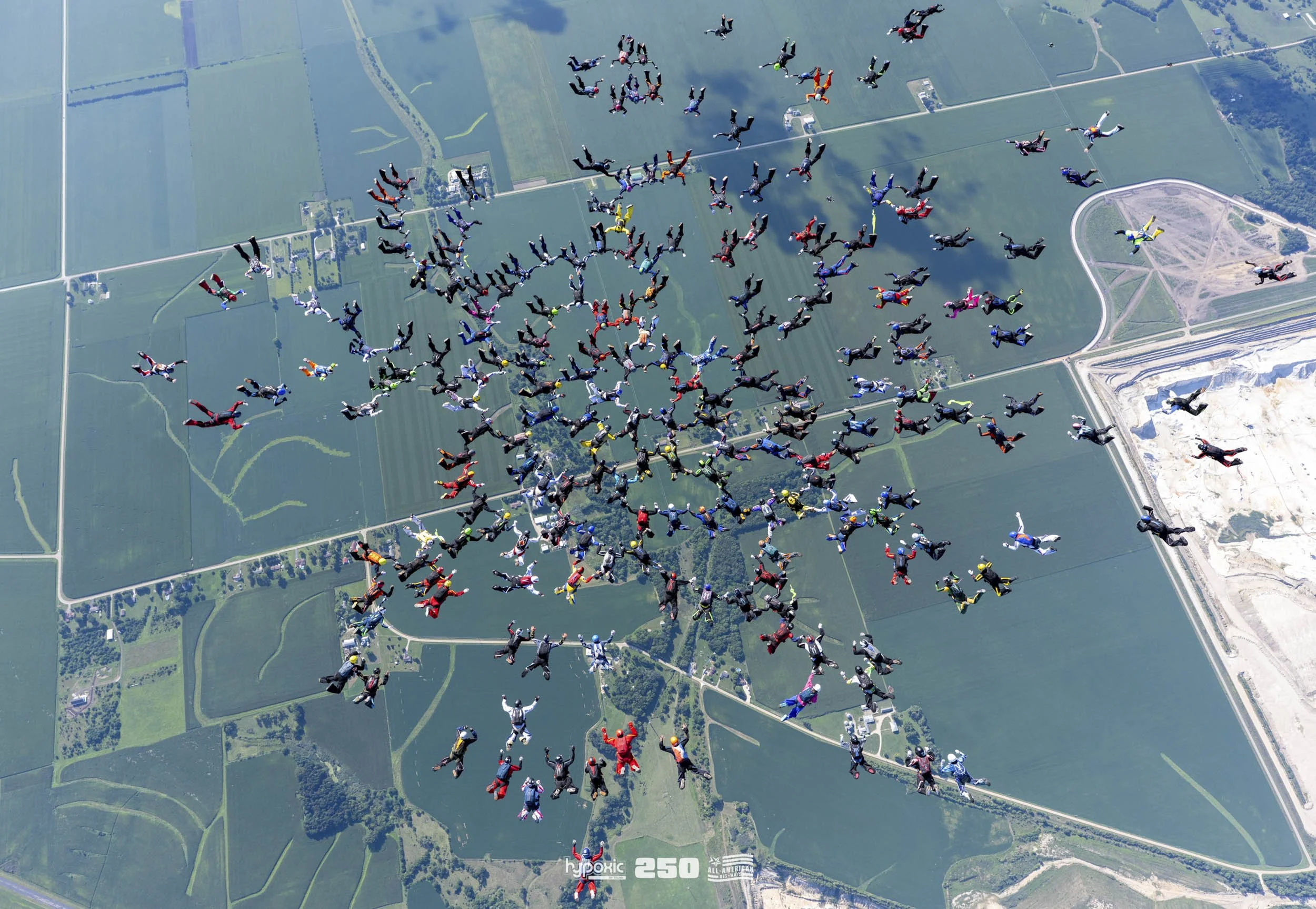 Skydivers in colorful jumpsuits and gear descending over open farmland, during a group jump, with fields and roads below.