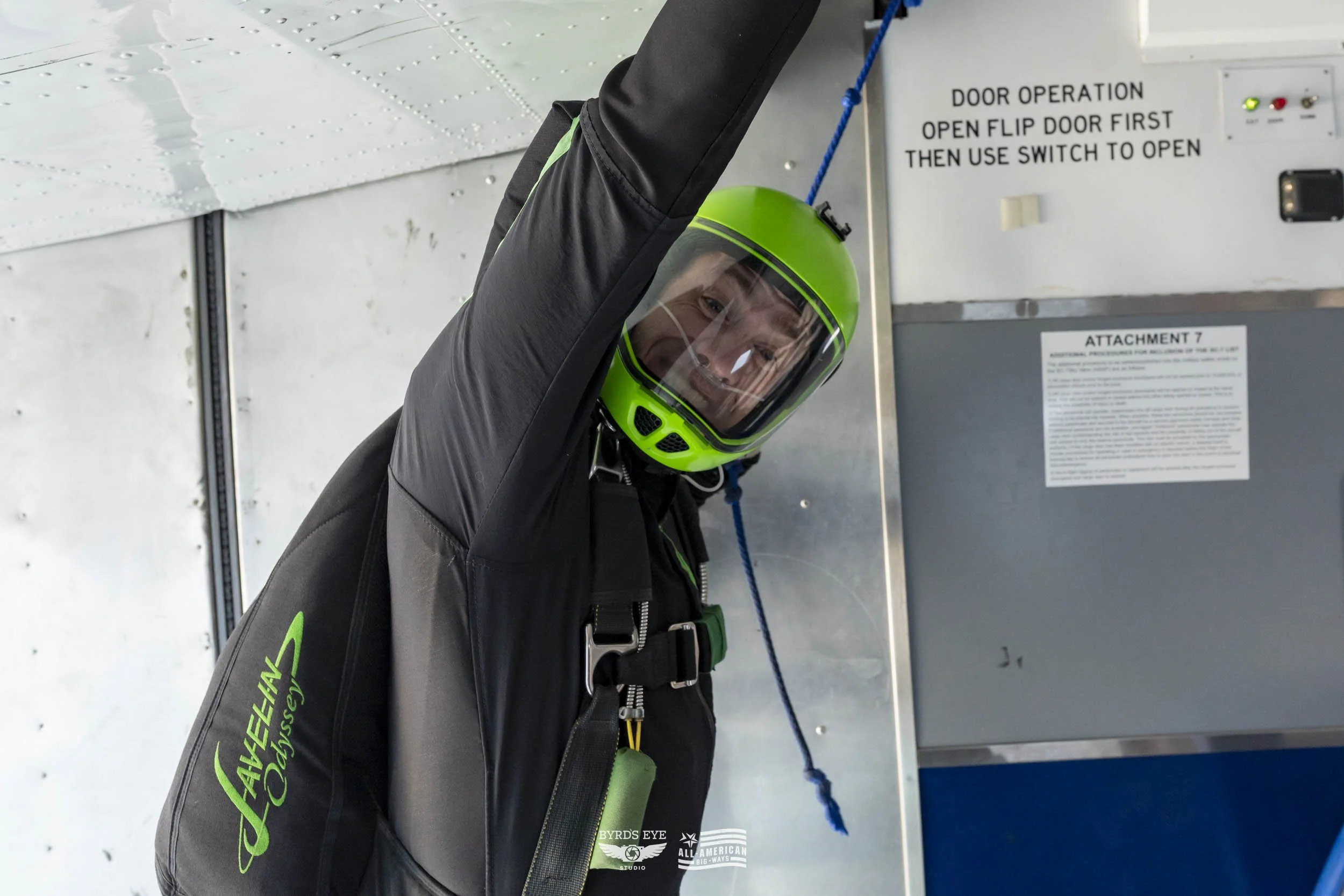 A person in a black and gray jumpsuit with 'Aviation Odyssey' written on the sleeve, wearing a bright green helmet and a transparent face shield, inside an aircraft preparing to skydive. The person is raising one arm and smiling.