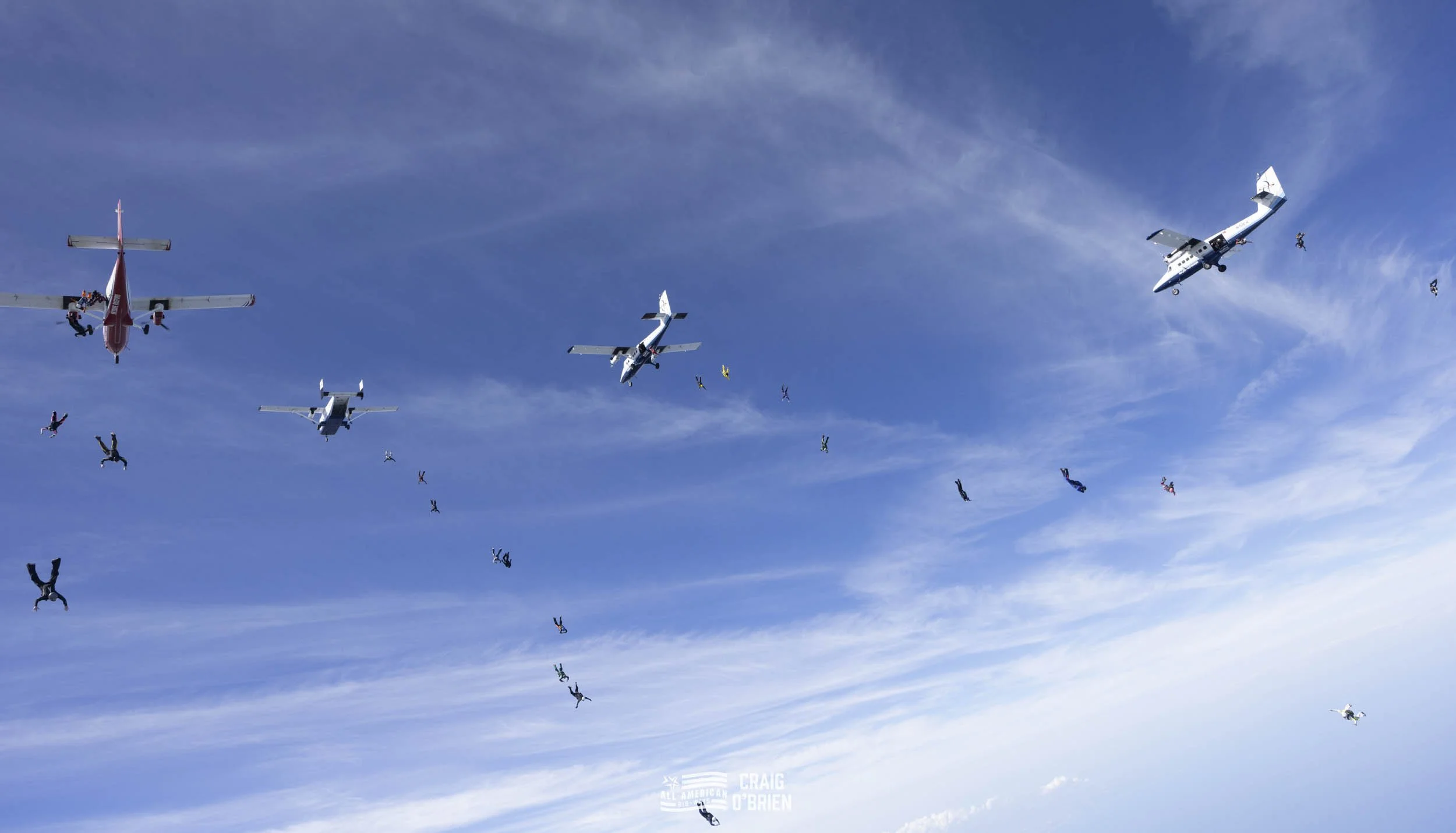 Multiple skydivers in free fall beneath several aircraft flying in a clear blue sky.