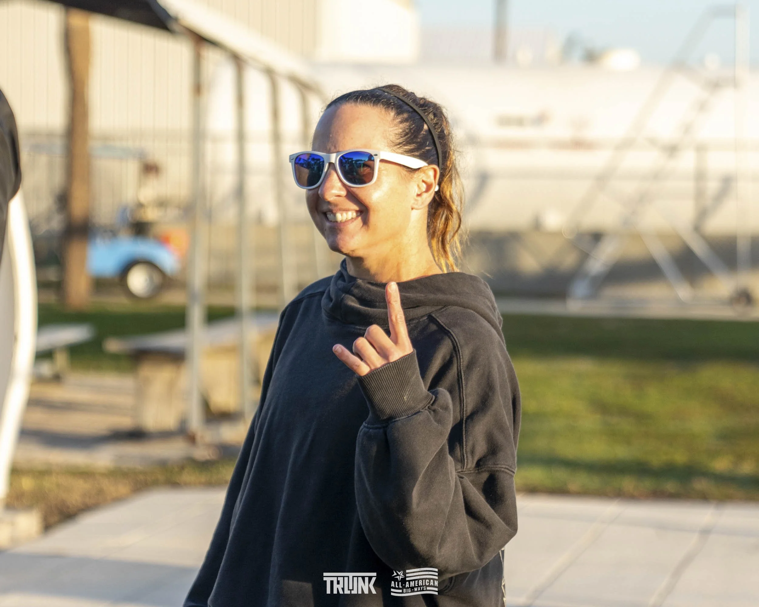 A woman wearing sunglasses smiling outside, pointing upward with her index finger. She has brown hair tied back, and is dressed in a black hoodie. In the background, there are boats and a fence.