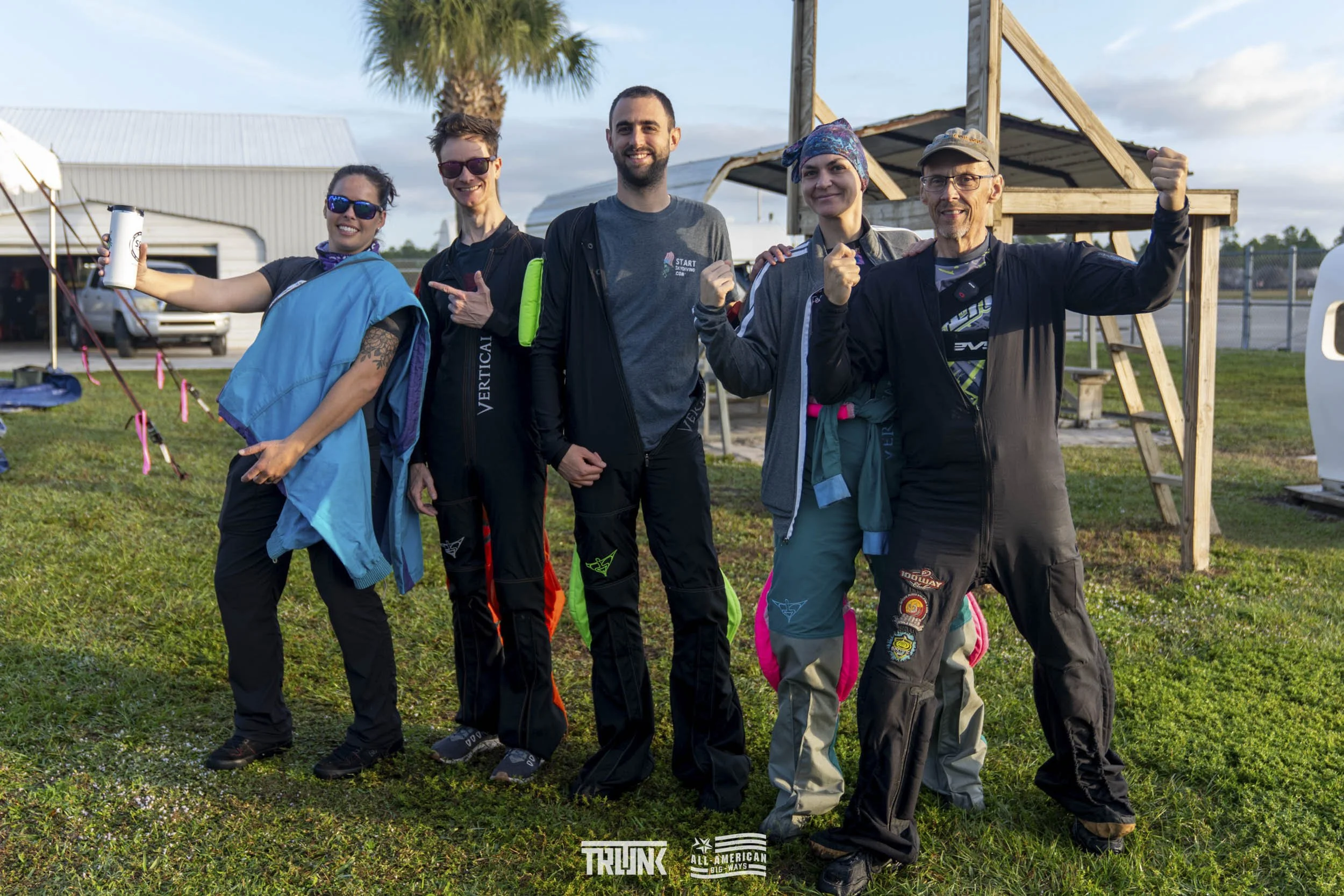 Group of six diverse people standing outdoors on grass, smiling, with some raising their fists or pointing, in front of a small wooden structure, on a partly cloudy day.