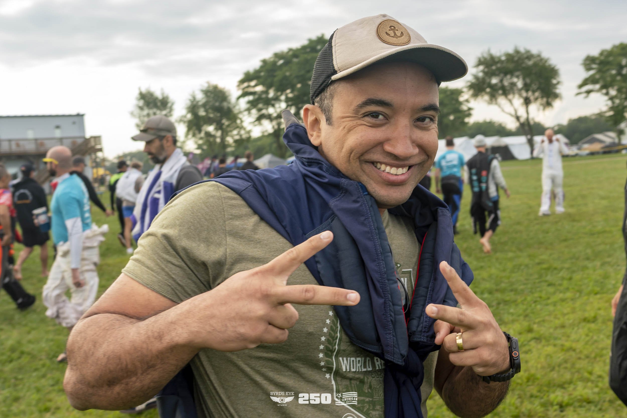 A smiling man wearing a beige baseball cap, khaki t-shirt, and a blue vest, making peace signs with both hands at an outdoor event with many people and tents in the background.