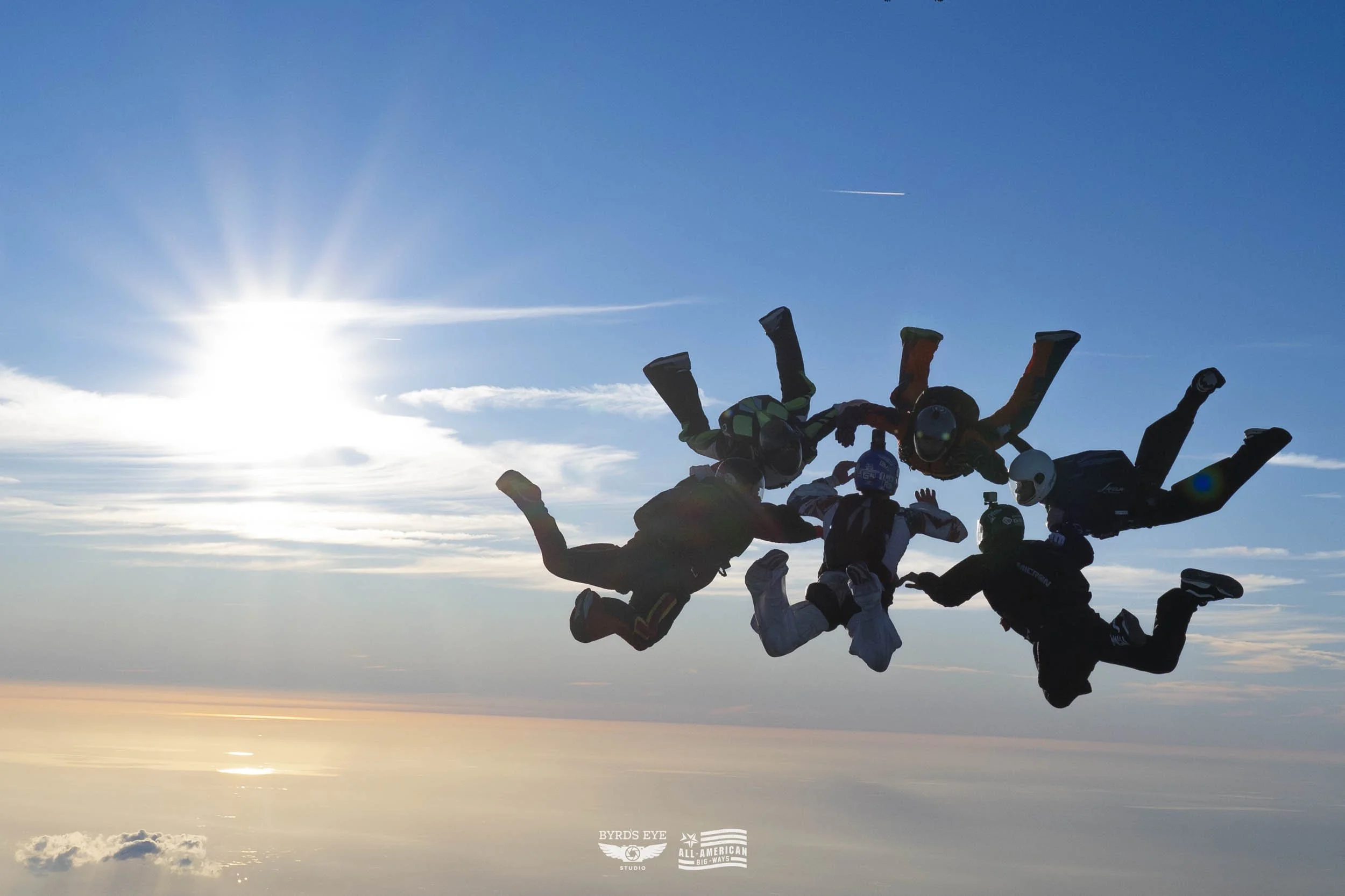 Five skydivers in freefall formation against a bright blue sky with the sun on the left and clouds below.