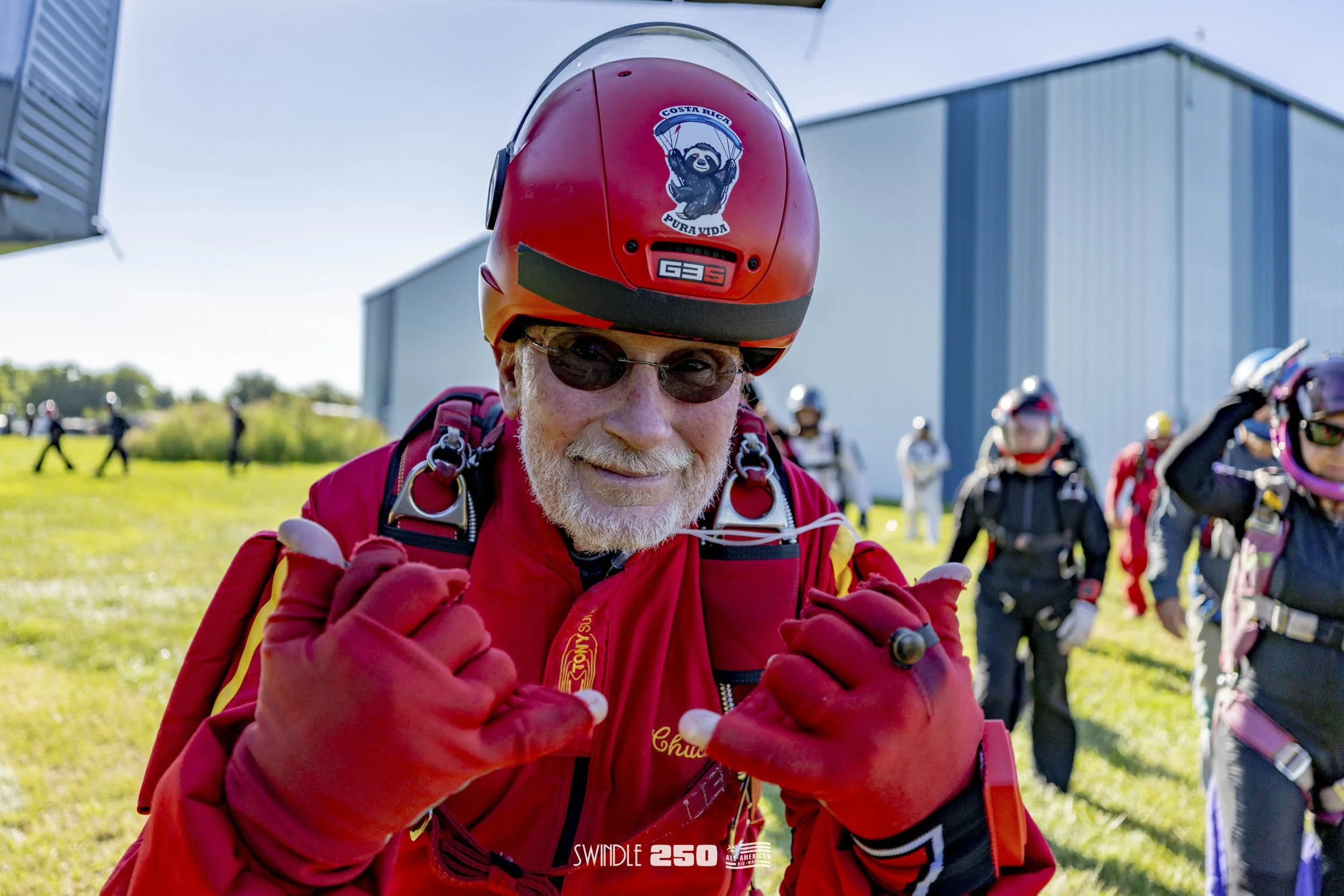 An older man with a beard and sunglasses smiling at the camera while wearing a red skydiving jumpsuit and helmet, with other skydivers in the background at a skydiving facility.