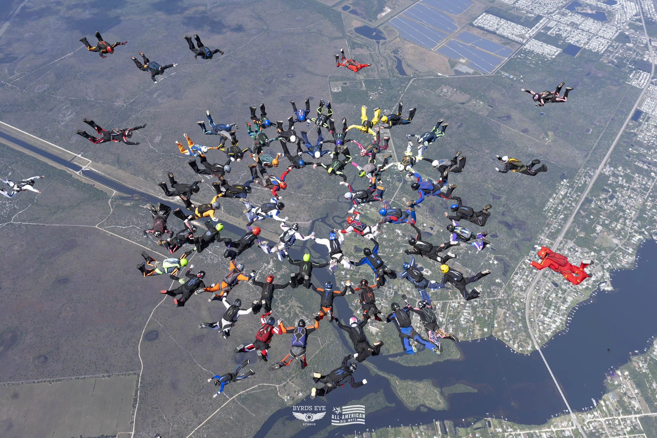 Group of skydivers forming a large human chain while free-falling over a landscape with water, roads, and fields.