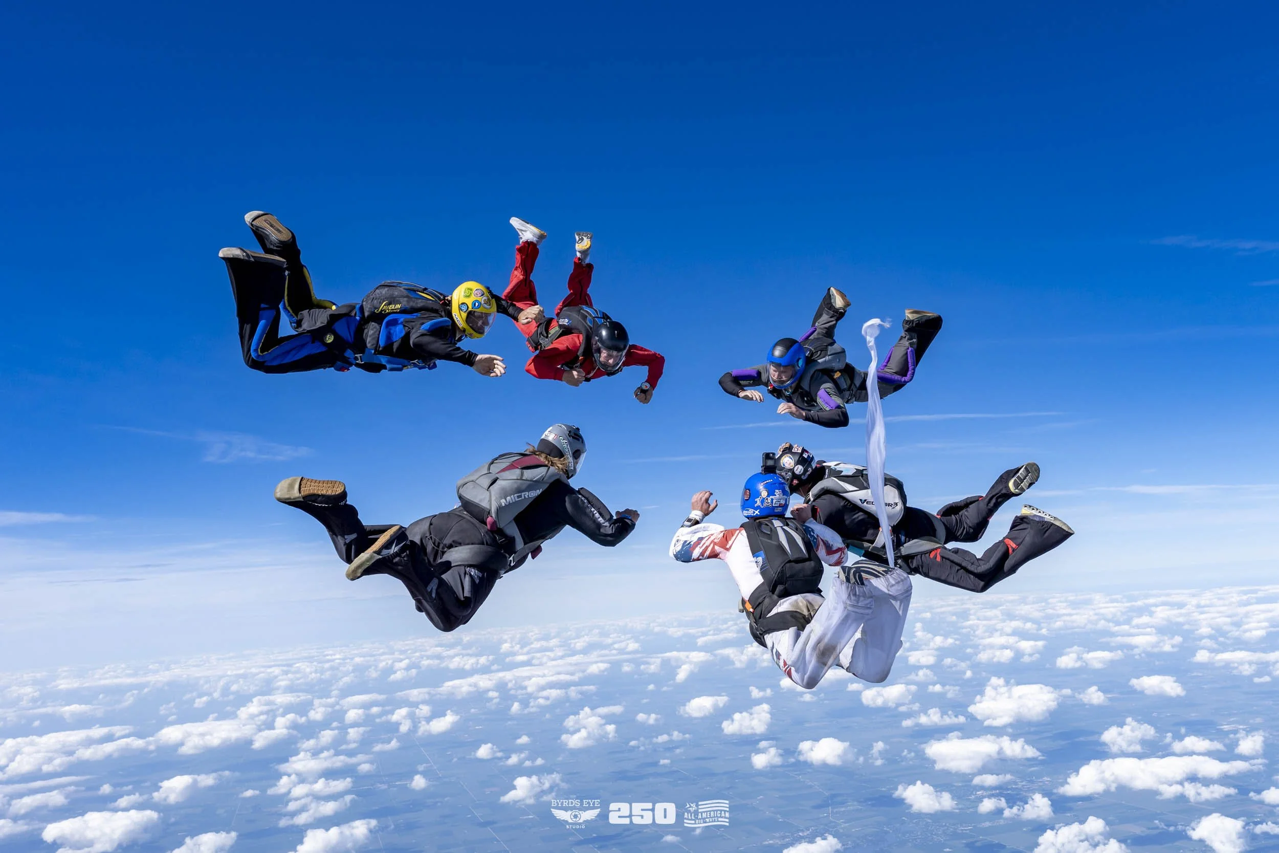 Group of skydivers in free fall above the clouds, wearing jumpsuits and helmets, forming a circle and holding hands.
