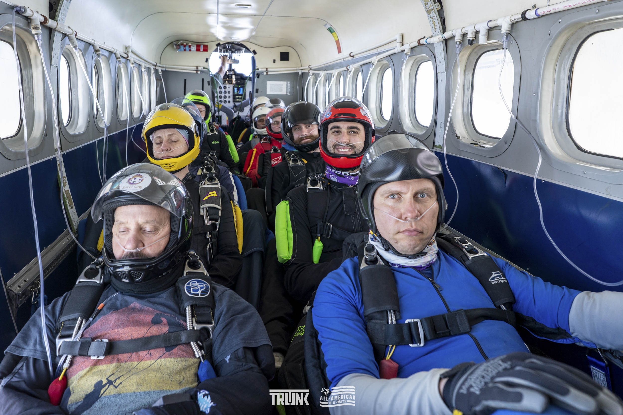 A group of skydivers sitting inside an aircraft, wearing helmets and jumpsuits, preparing for a jump.