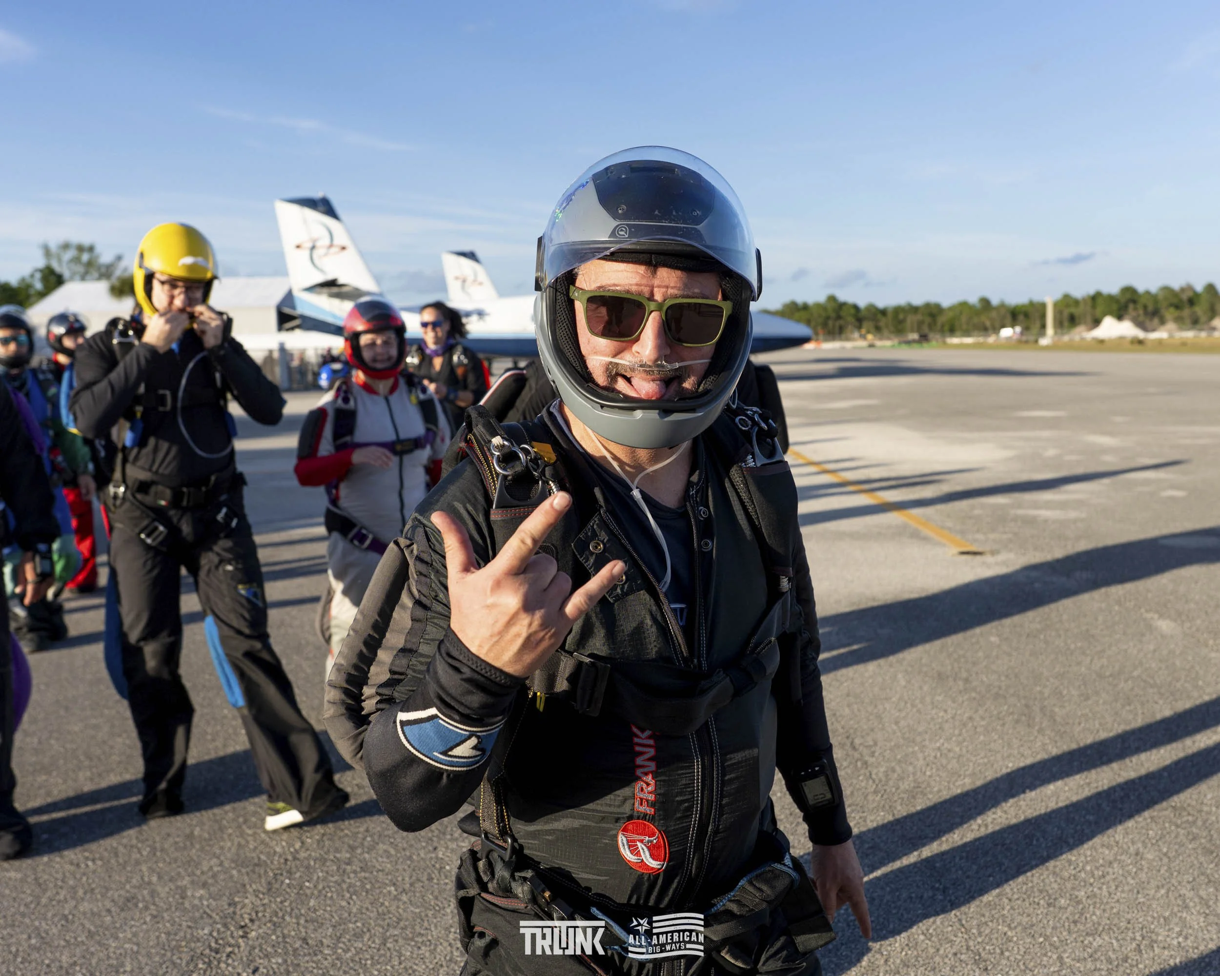 Parachutists preparing for a skydiving jump on the tarmac, with planes in the background and clear sky.
