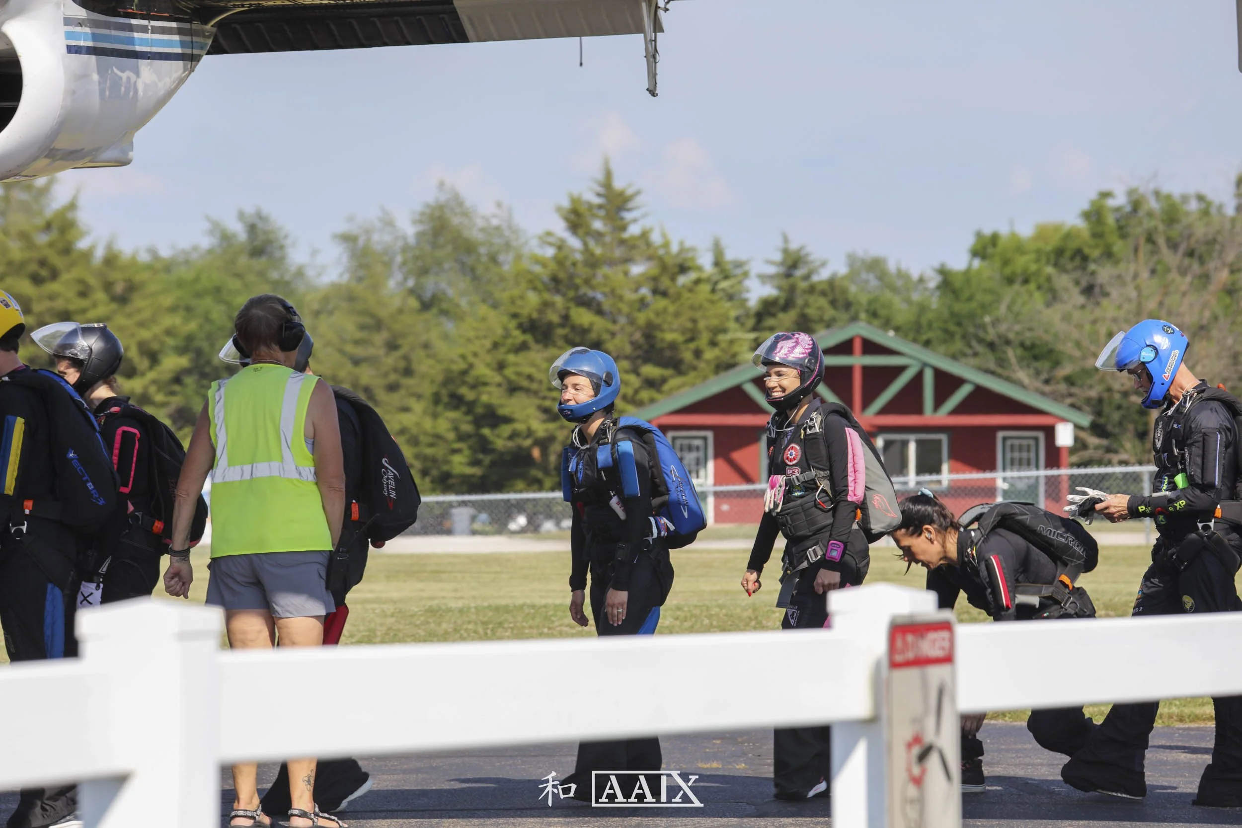 Group of skydivers in gear standing on grass, with a woman in a sleeveless neon vest and gray shorts facing away from the camera. A small red building and trees are in the background on a clear day.