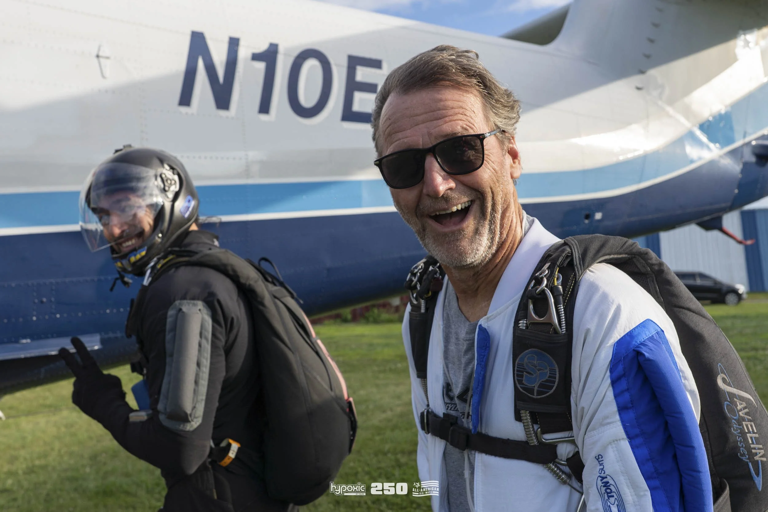 Two skydivers standing in front of a white and blue airplane, smiling and making peace signs. The man in the foreground is wearing a white and blue jumpsuit, black sunglasses, and a black harness. The person in the background is wearing a black jumps