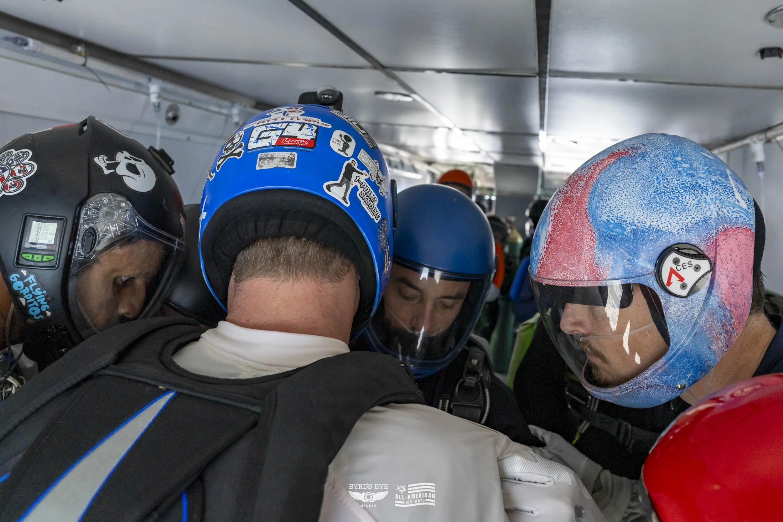 Group of skydivers in a huddle inside an aircraft, wearing helmets and jumpsuits, preparing for a leap.