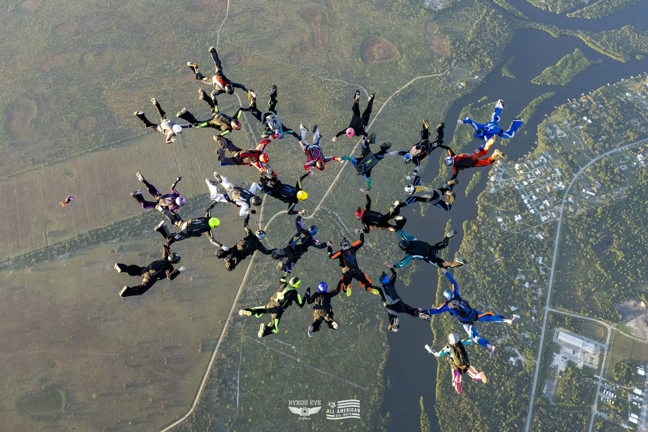 Skydivers in colorful jumpsuits and helmets forming a star shape in mid-air above a landscape with water, trees, and roads.