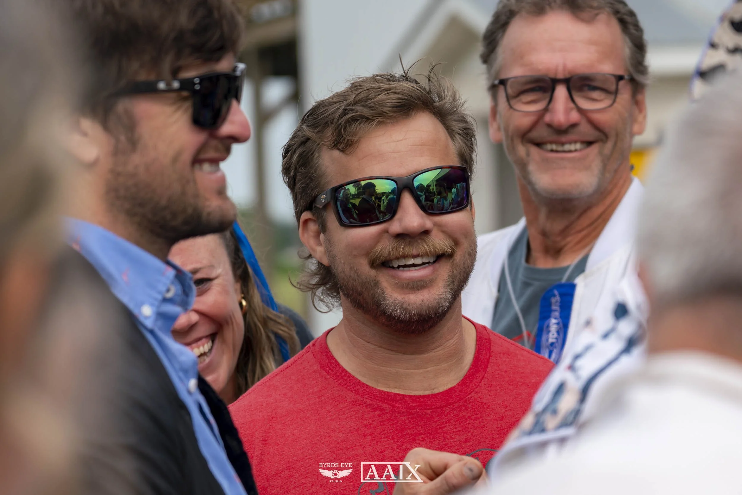 Group of people smiling and laughing, wearing sunglasses, one person in a red shirt, outdoors in a cheerful setting.
