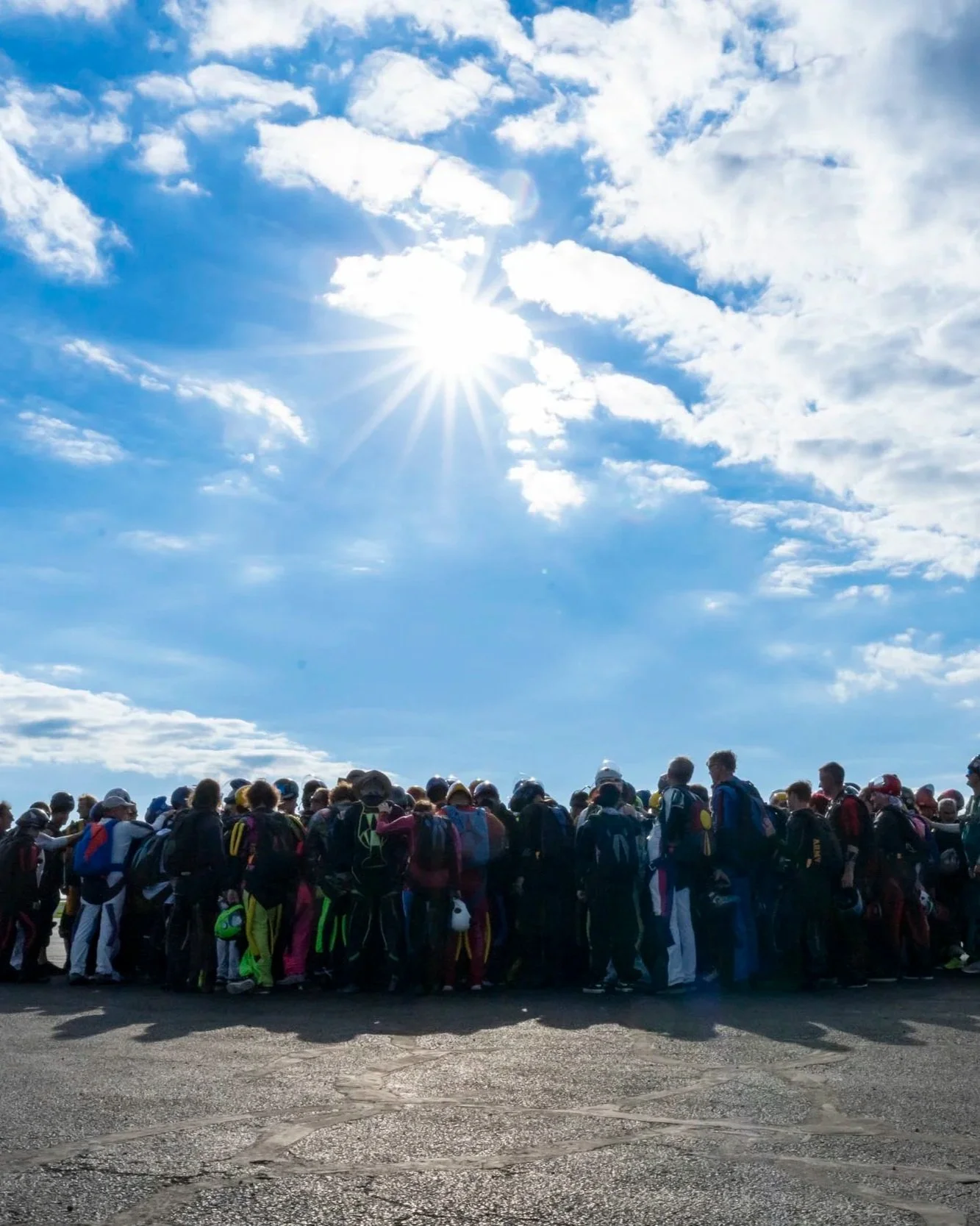 A large group of people gathered outdoors under a bright blue sky with scattered clouds and the sun shining brightly.