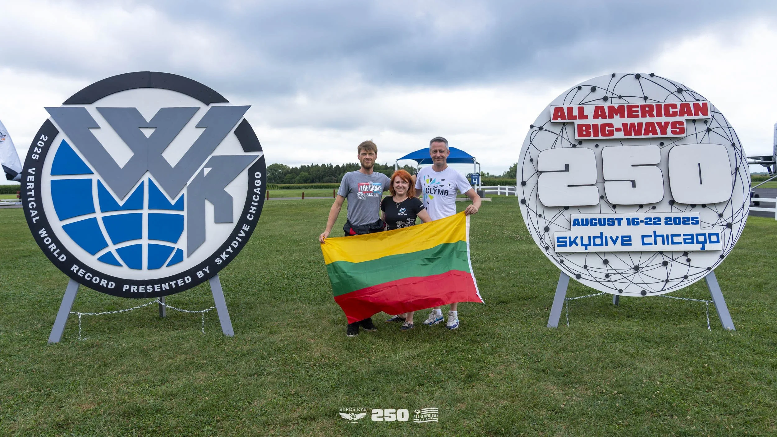Three people standing on a grassy field holding a Lithuanian flag, with skydiving event signs on either side. The left sign features a logo for the World Record presented by Skydiving Chicago, and the right sign indicates an all-American big-ways for