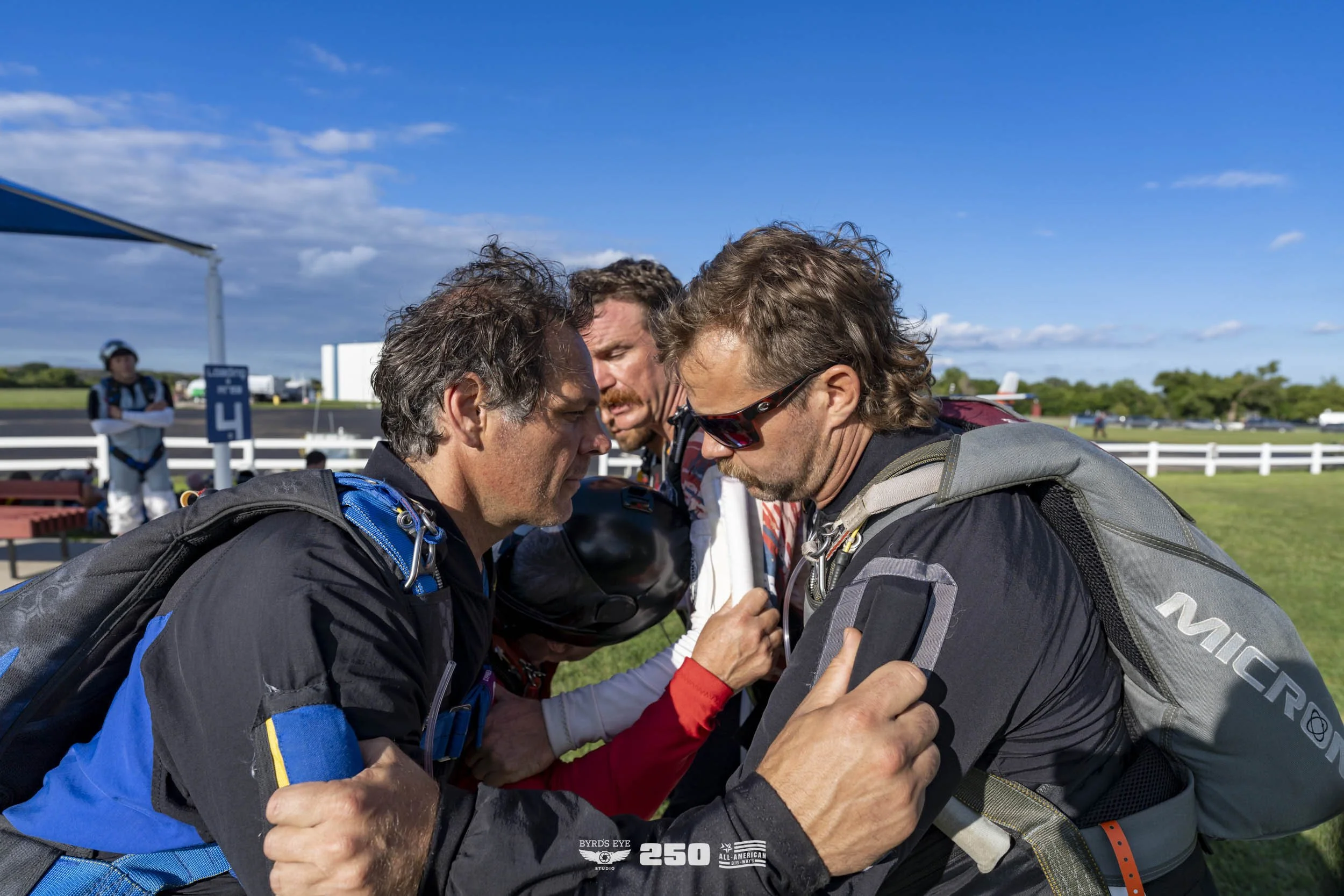 Skydivers in a huddle on a grassy field preparing for jump, with clear blue sky and white fence in background.