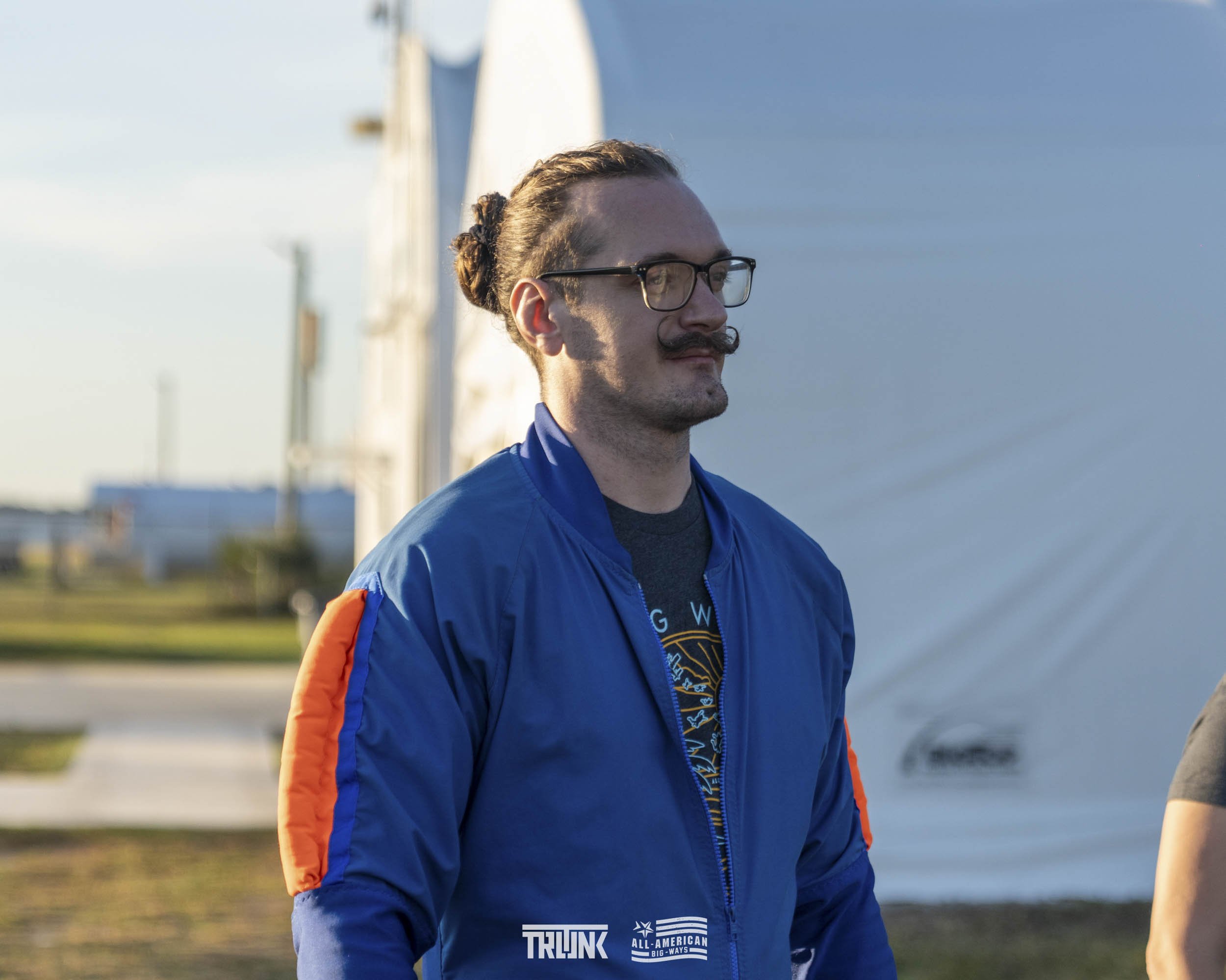 A man with glasses and a mustache, wearing a blue jacket with orange stripes on the sleeves, standing outdoors with a large white structure and a grassy area in the background.