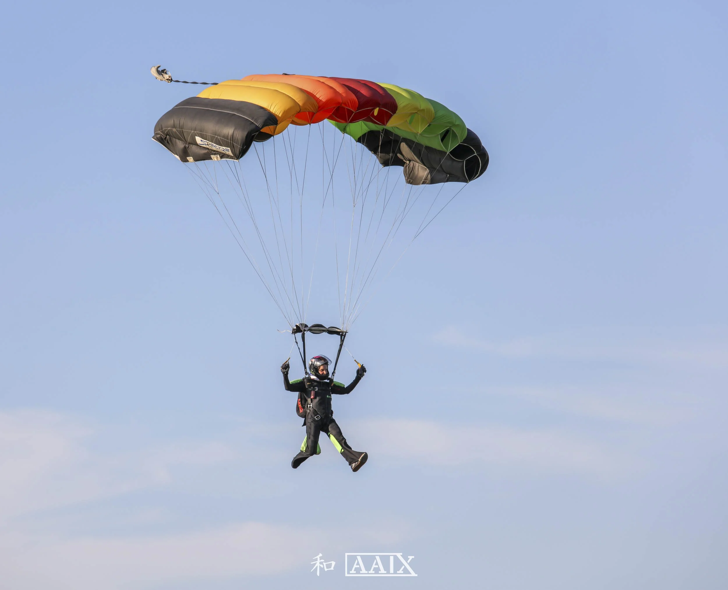 Person in skydiving gear descending with a colorful parachute against a clear blue sky.