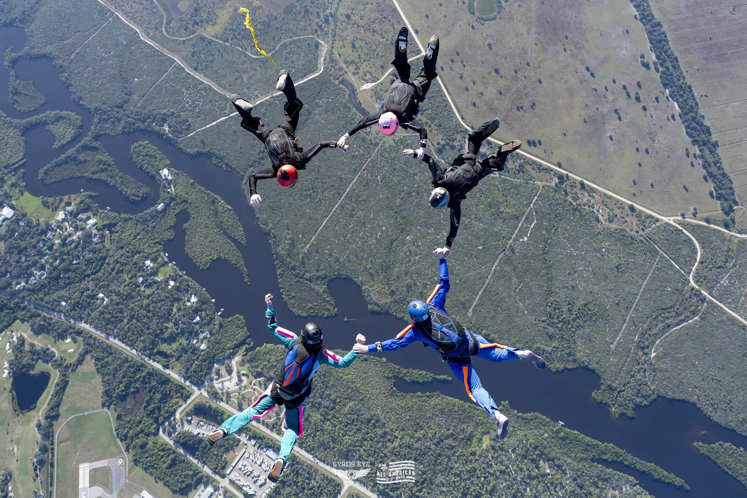 Six skydivers holding hands in a circle while free-falling above a landscape with lakes, trees, and roads.