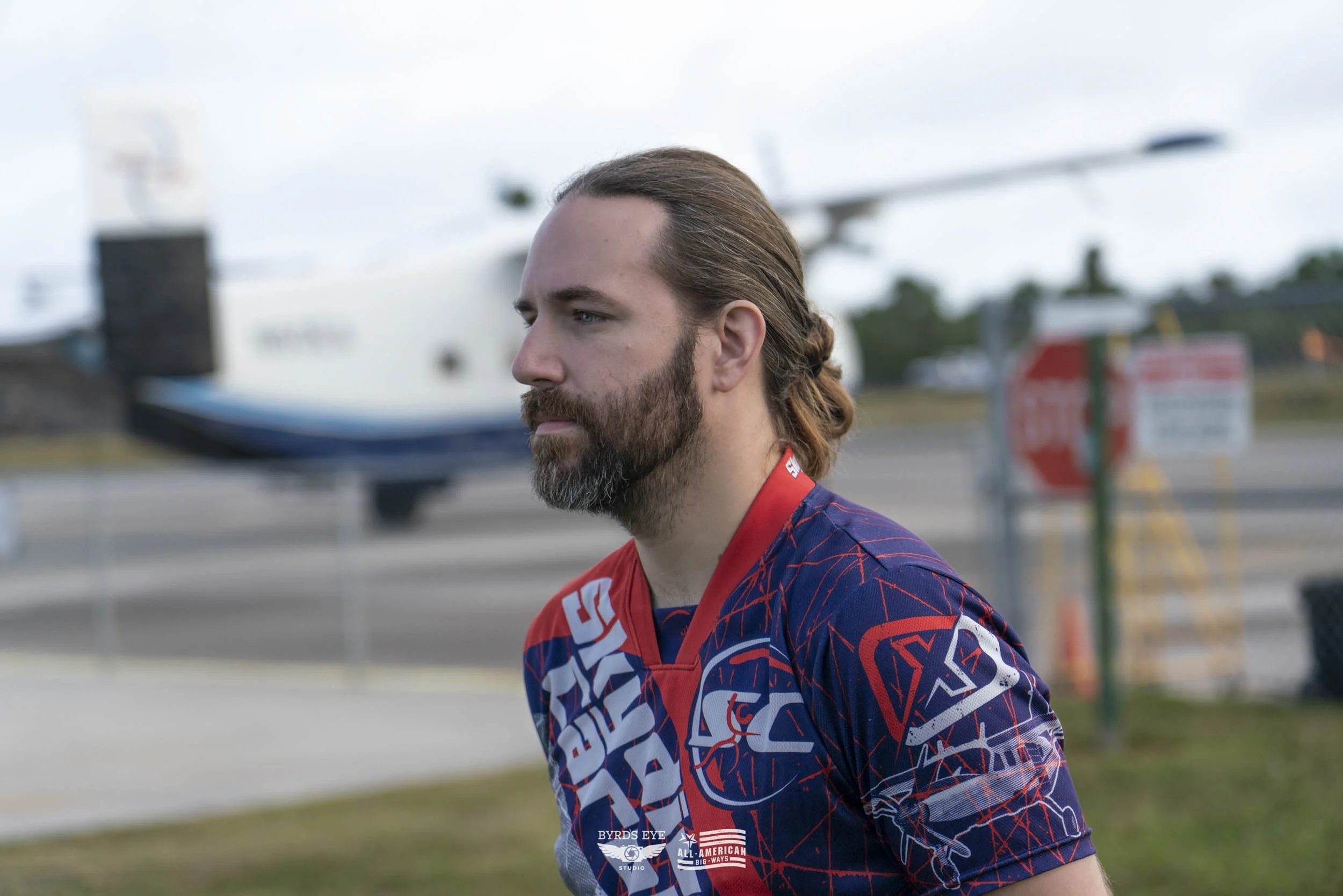 A man with long hair and a beard wearing a sports jersey stands outdoors near an airport tarmac with a large aircraft in the background.