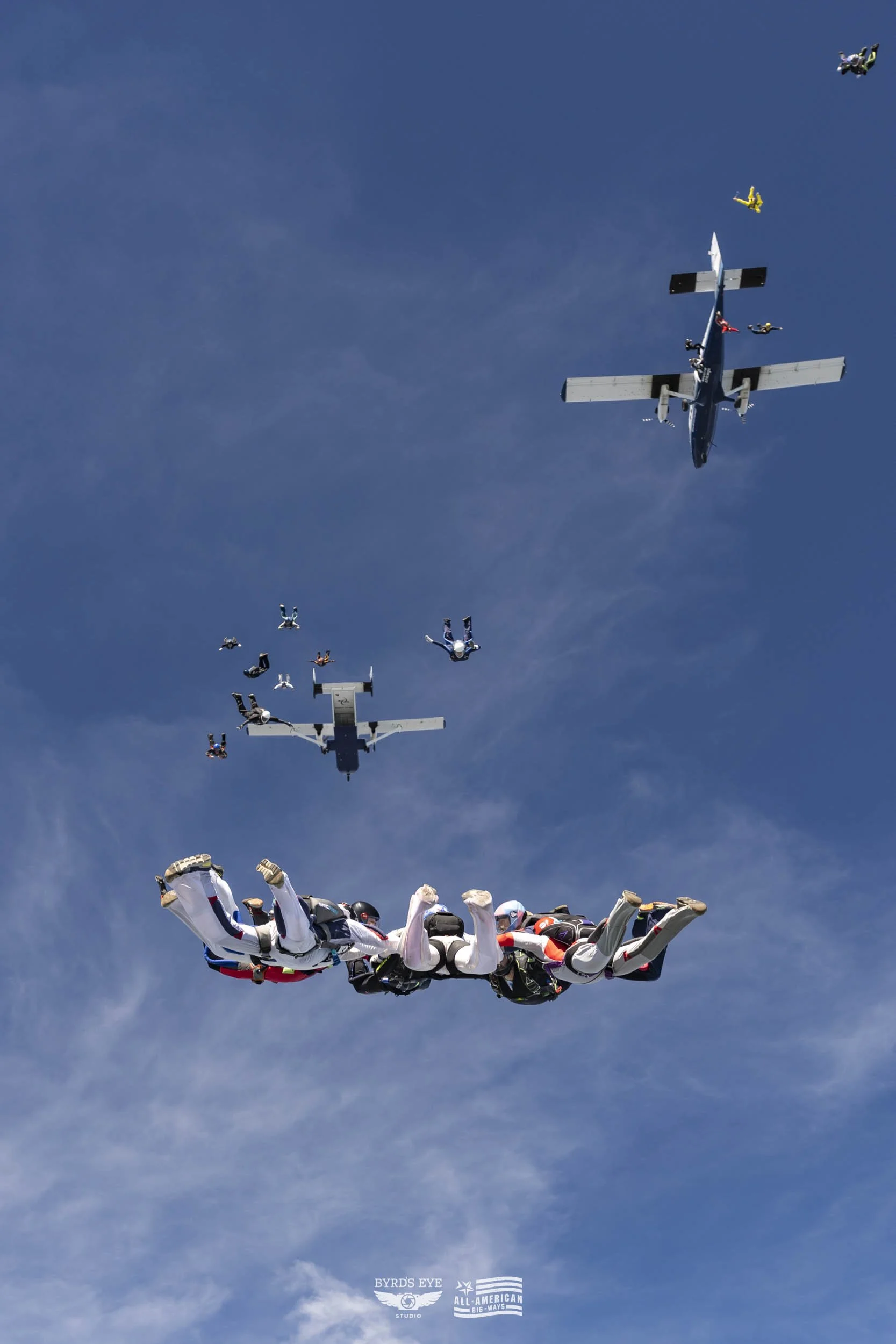 Skydivers in free fall forming a group, with two planes above them in a clear blue sky.