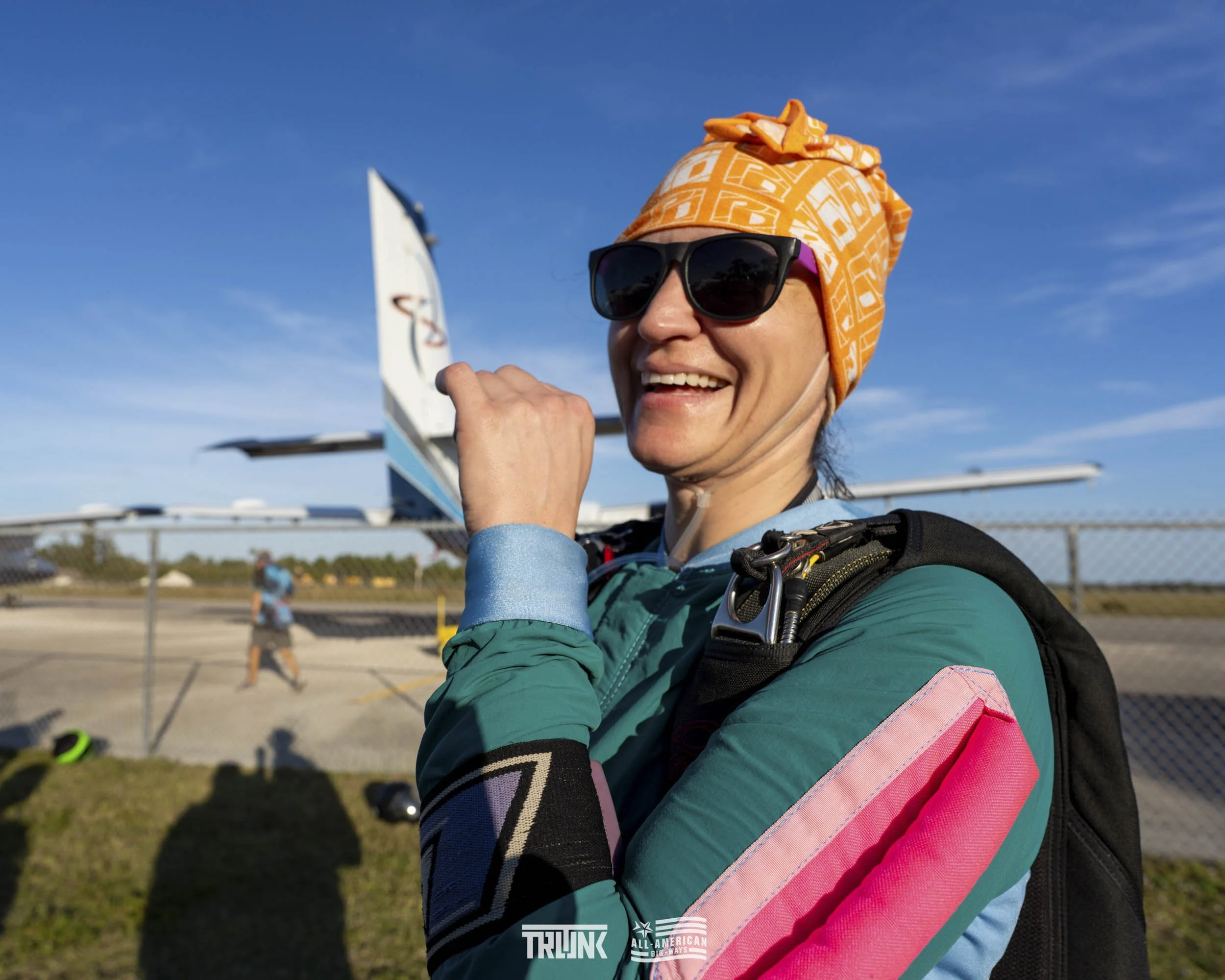 A woman in skydiving gear, wearing sunglasses and an orange headwrap, smiling at the camera with a small aircraft in the background on a clear day.
