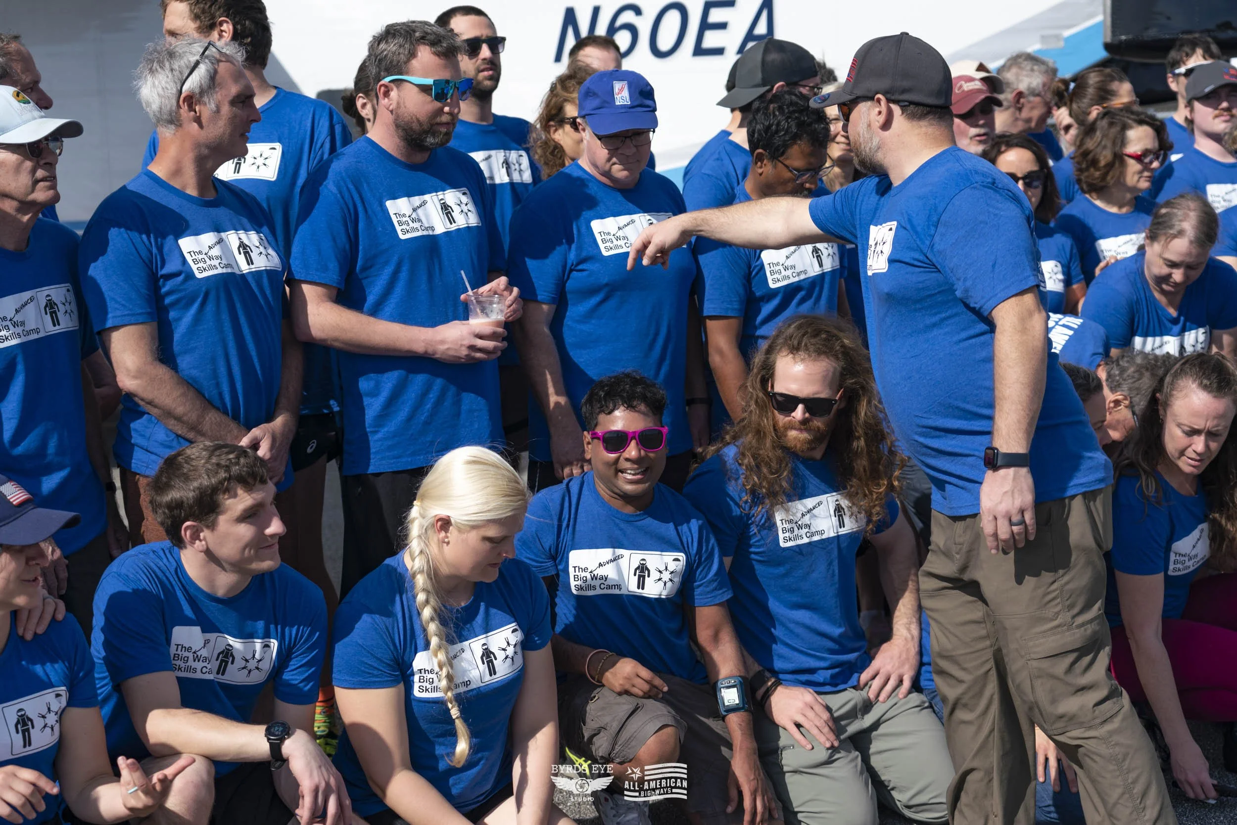 A group of people wearing blue T-shirts with 'The Big Way Skills Camp' logo, gathered outdoors, some sitting and some standing, with a man in front of them pointing, possibly during a group event or team activity.