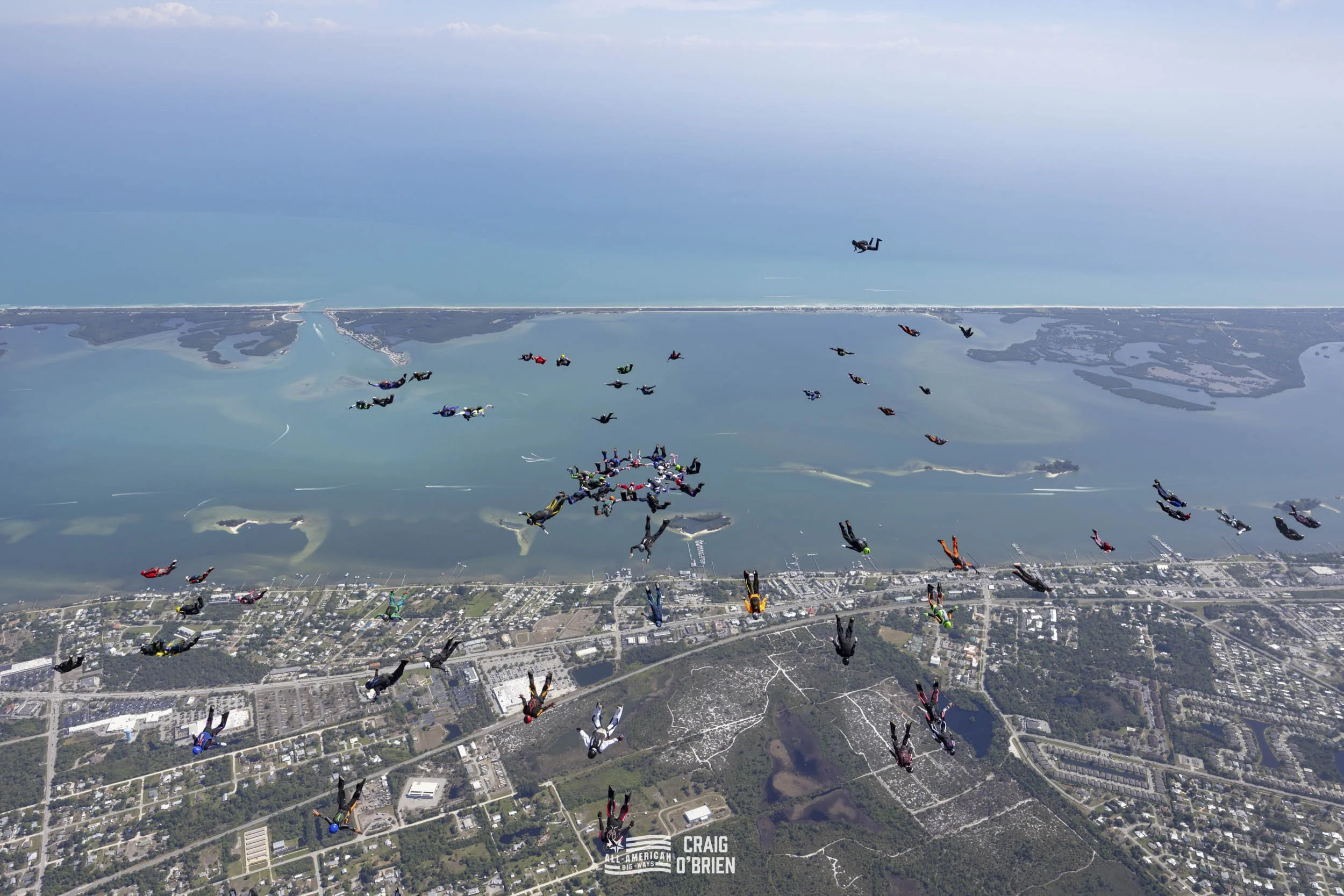 Group of skydivers in freefall formation over a landscape with water and land, during daytime.