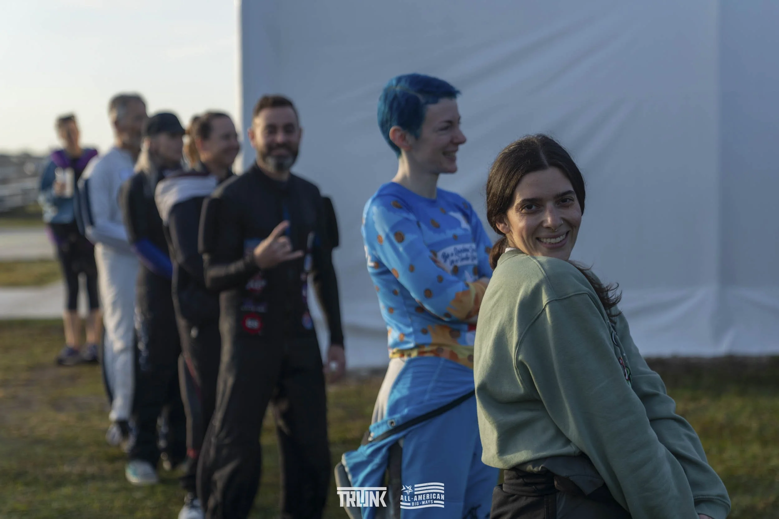 Group of people standing in a line outdoors, smiling, with some making hand gestures, in front of a white structure.